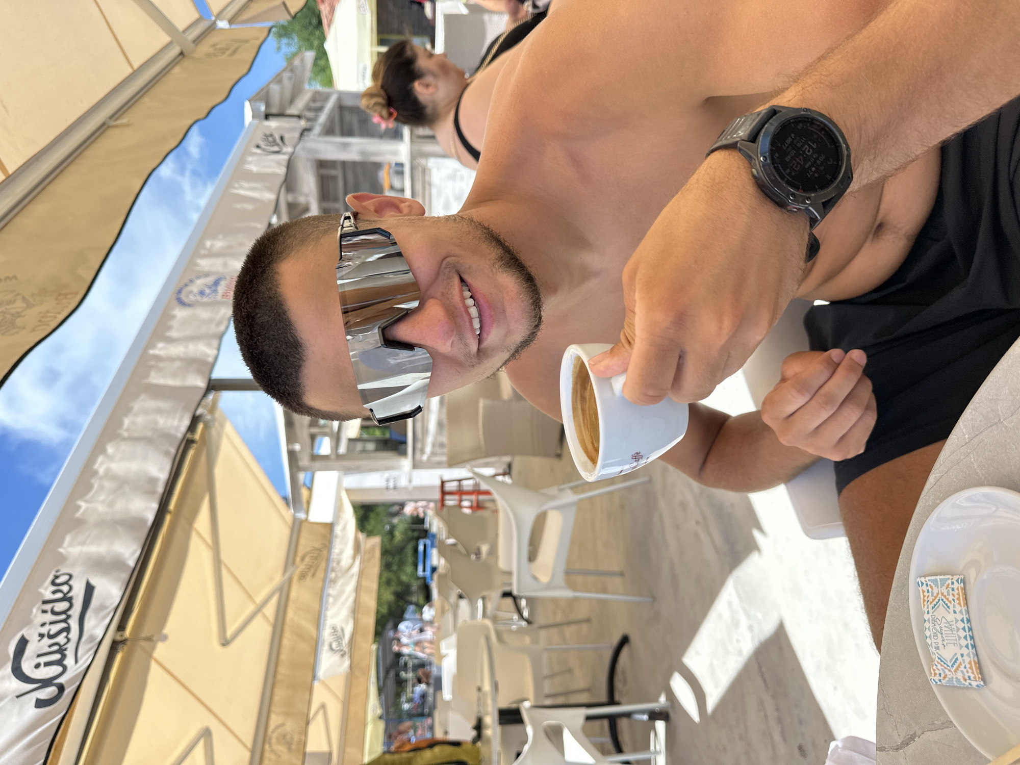 A man, facing the camera, is holding a small white cup of coffee. He is wearing mirrored, wraparound sunglasses and a dark-colored watch on his left wrist. He has a short beard and is smiling. The man is shirtless, and appears to be seated at a table in an outdoor cafe. Behind him, another person is visible, also wearing sunglasses. The cafe appears to be set up with tables and chairs, some of which are white plastic. Tented awnings are overhead, suggesting a sunny day. The man's watch displays the time as "12:42".
In the background, other patrons are seated at tables, and there are glimpses of foliage and beach umbrellas. The overall scene suggests a relaxed atmosphere at a beachside cafe. Visible text on the awnings reads "Čikšidko". A small sugar packet on a white saucer near the man reads "WHITE Sugar Diamond Pure". The location is identified as Budva, Montenegro. The time of day appears to be around midday. The weather seems fair and sunny.