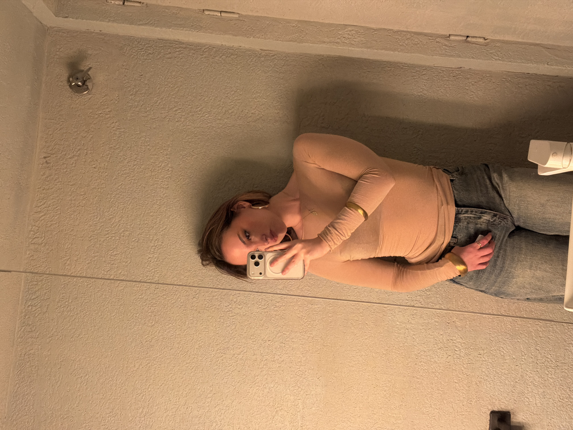 A woman stands in a bathroom, taking a selfie in the mirror. She is wearing a long-sleeved beige top, blue jeans, and gold jewelry. The bathroom has a textured wall, a light fixture, and a toilet paper dispenser. The location is not discernible from the image provided.