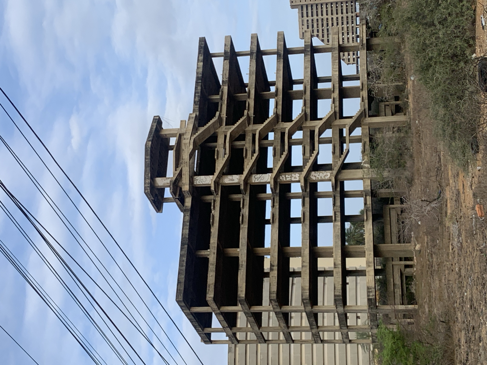 An unfinished or abandoned concrete building structure dominates the central and right portions of the image, standing upright with a skeletal framework of pillars and horizontal slabs. Its distinctive design features multiple projecting, angular elements resembling stairs or cantilevered balconies, forming a complex, open grid pattern. Visible graffiti tags, including "SWO" and "CRWS," are present on some of the lower concrete beams.
To the right and at the base of the structure, the ground is dry and covered with sparse, scrubby vegetation, alongside some larger bushes and small trees. In the background, behind the skeletal building, a taller, completed building with numerous rectangular windows or facade elements is visible. The sky, seen through and above the concrete frame and on the left side of the image, is partly cloudy with patches of clear blue. Several dark utility wires run diagonally across the upper left portion of the sky.
The scene depicts an urban or peri-urban environment in Porlamar, Venezuela, during the daytime, likely under bright, natural light from mid-morning or afternoon. There is no human activity visible, conveying a sense of halted development or abandonment.