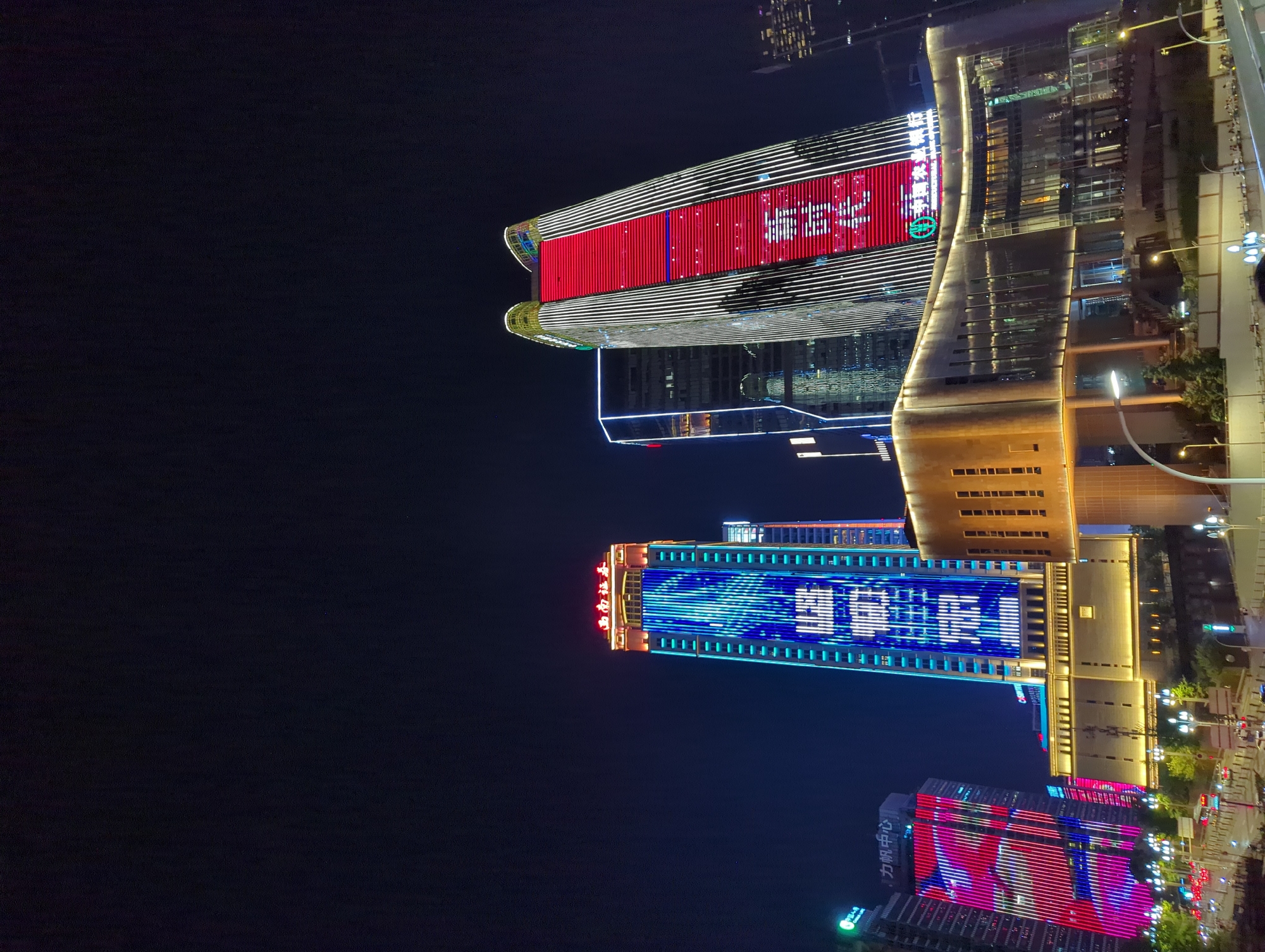 The image captures a nighttime view of a city skyline, specifically the area of 江北城街道, China, dominated by several illuminated skyscrapers. The tallest building on the left displays a vertical pattern of blue and white lights forming Chinese characters. To its right, another skyscraper is clad in vibrant red and white vertical lighting, also featuring Chinese characters. In front of these tall structures, a more modern, low-rise building with a curved facade is brightly lit with warm yellow lights, showcasing a glass-fronted interior. The surrounding area includes streetlights, trees, and a street with moving traffic, suggesting an active urban environment. The sky is dark, indicating it is nighttime, and the overall impression is one of a brightly lit, modern cityscape.