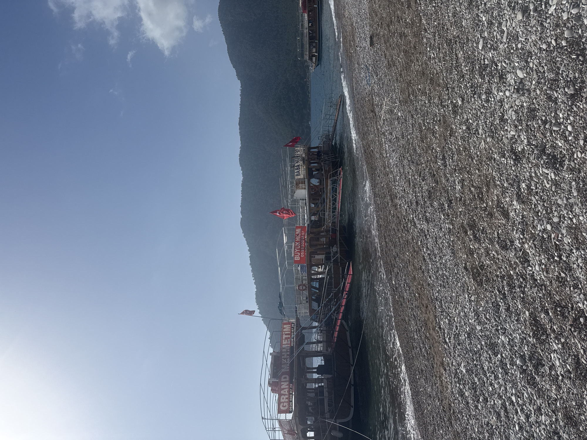 The image captures a beach scene in Kumluca, Turkey, where several tour boats are docked along the shoreline. The boats, identified by signs reading "GRAND MEHMETIM", "BUYUK HILMI," and "BABA NURI 37," suggest a tourism-related activity. A pebble-strewn beach stretches along the foreground, meeting with gentle waves. In the background, a mountain ascends into a clear blue sky with light clouds. The scene is bathed in sunlight, implying a warm, pleasant day.