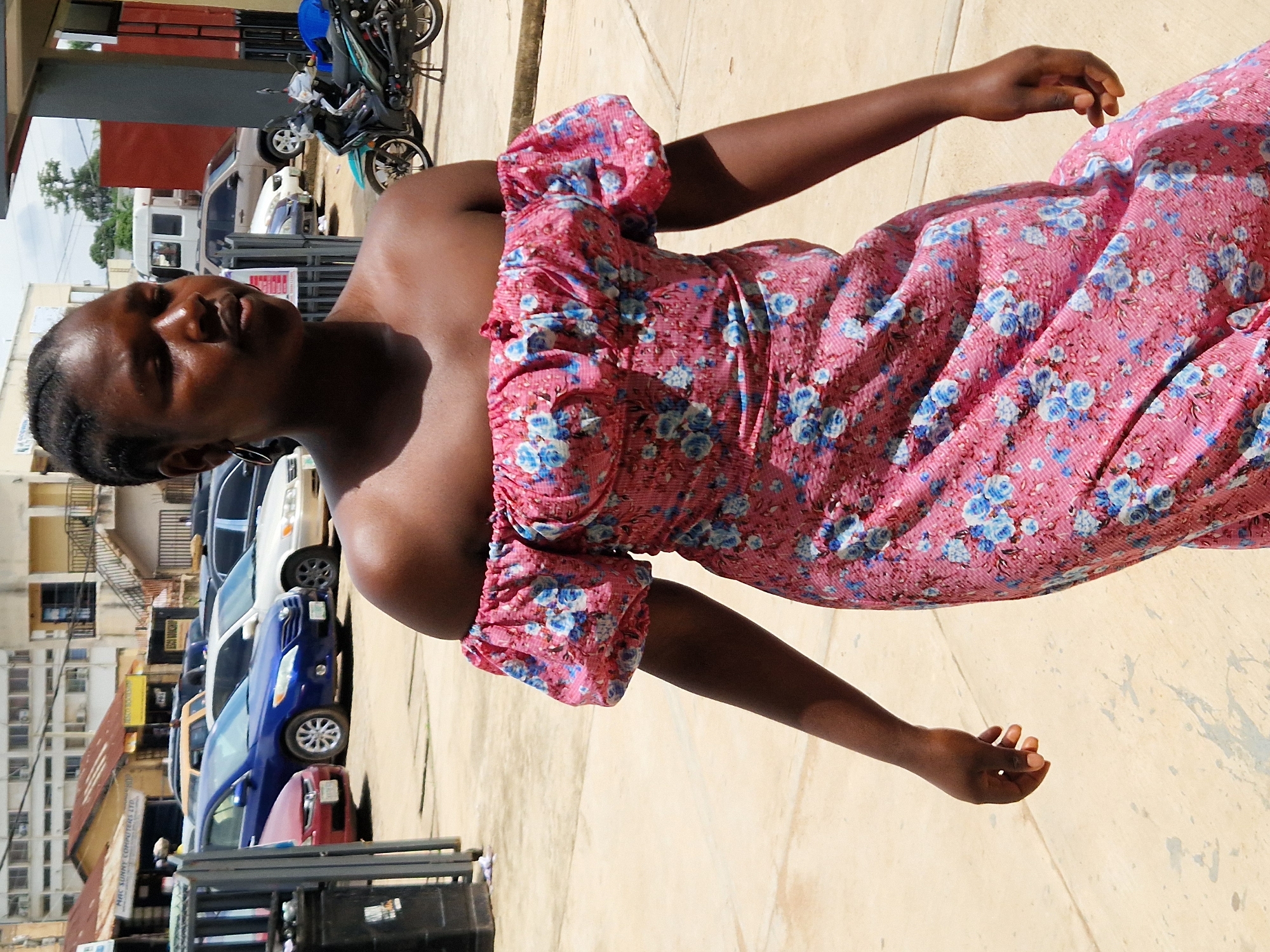 A young woman with dark skin and braided hair, wearing an off-the-shoulder pink floral dress, walks outdoors on a paved surface. Her head is turned slightly to her right, and her eyes are looking down. Her right arm is bent at the elbow, with her hand relaxed at her side. Her left arm is also bent, with her hand extended downwards, fingers slightly curled. In the background, several cars are parked, including a blue sedan and a white minivan. A motorcycle with a black and turquoise body is parked to the right of the woman. Buildings are visible in the background, including a multi-story structure with windows. The overall scene appears to be a street or parking area in Jos, Nigeria, with the bright daylight indicating it is daytime.