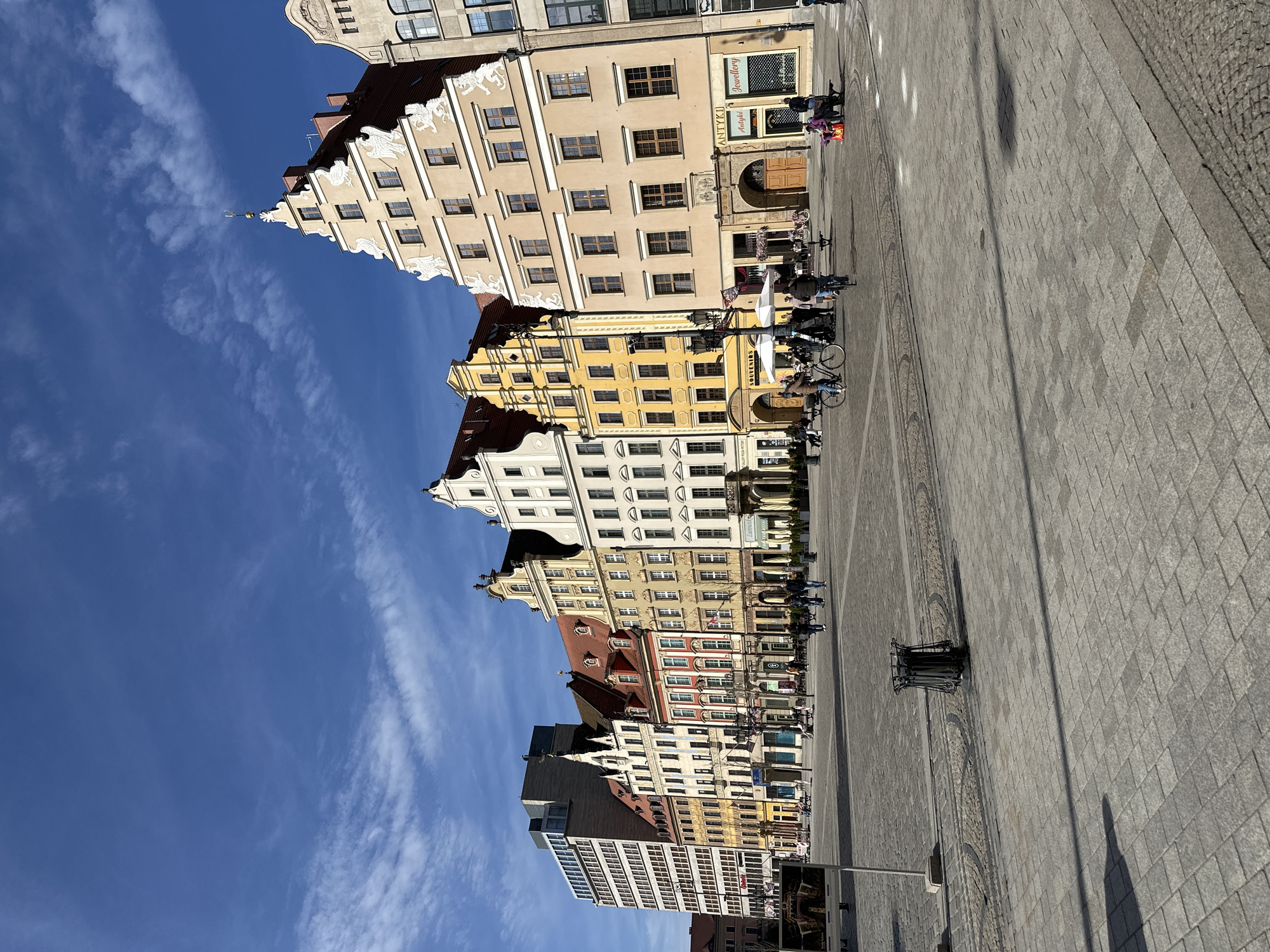 The image shows a view of the Market Square in Wrocław, Poland. The square is paved with gray stone and surrounded by colorful historic buildings. People are visible walking and sitting outside cafes along the buildings. The sky is blue with light clouds.