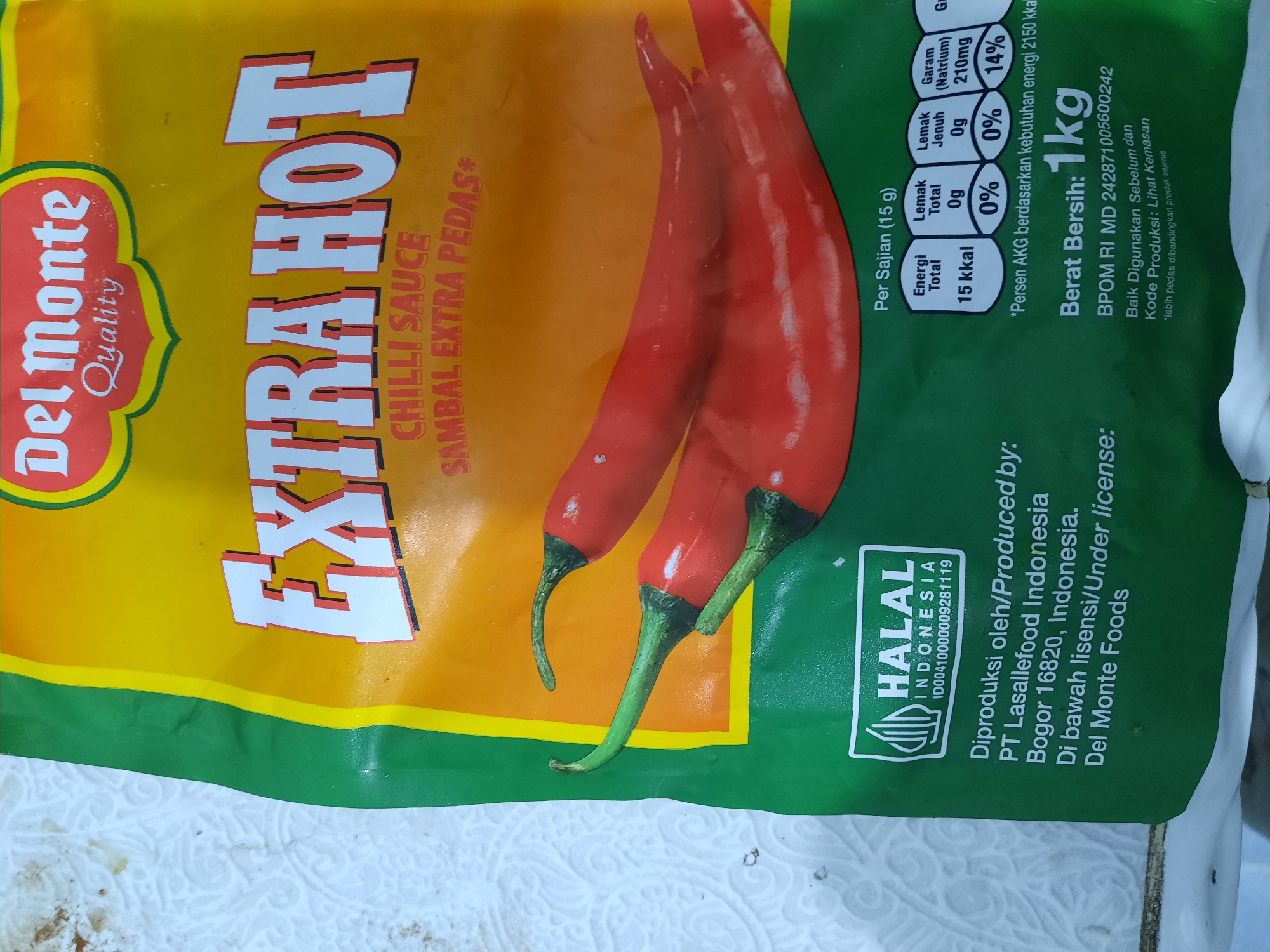 This is a close-up, vertically oriented photograph of the packaging for Del Monte Extra Hot Chilli Sauce. The dominant colors are yellow, orange, and green, with bright red chili peppers prominently featured on the orange section of the packaging.
The subjects are the product packaging itself, designed to attract consumers with its bold "EXTRA HOT" lettering in large white font with a dark outline. The branding "Del Monte Quality" is visible on the left side. Two red chili peppers are depicted, suggesting the spiciness of the product.
There are no people or animals in the image. The scene is the product packaging, likely photographed in a domestic or retail setting, with a white textured surface visible at the bottom, possibly a paper towel or a patterned tablecloth, which could be in Lhokseumawe, Indonesia, as per the location context.
Several details are visible in text on the packaging:
- "EXTRA HOT CHILLI SAUCE SAMBAL EXTRA PEDAS*"
- Nutritional information is presented in a table, including "Per Sajian (15 g)", "Energi Total 15 kkal", "Lemak Total Og 0%", "Lemak Jenuh Og 0%", and "Garam (Natrium) 210mg 14%".
- A statement about "Persen AKG berdasarkan kebutuhan energi 2150 kka" (Percentage of RDI based on energy needs of 2150 kcal).
- "Berat Bersih: 1kg" (Net Weight: 1kg).
- "BPOM RI MD 242871005600242".
- Instructions like "Baik Digunakan Sebelum dan Kode Produksi: Lihat Kemasan" (Best Before and Production Code: See Packaging).
- A note: "'lebih pedas dibandingkan produk sejenis" (spicier than similar products).
- A "HALAL INDONESIA" certification mark with a number "ID00410000009281119".
- Production details: "Diproduksi oleh/Produced by: PT Lasallefood Indonesia Bogor 16820, Indonesia. Di bawah lisensi/Under license: Del Monte Foods".
The image does not convey any specific time of day, weather, or emotions, other than the implied intensity suggested by "Extra Hot" and the chili peppers. The location context provided suggests it was photographed in Lhokseumawe, Indonesia.
