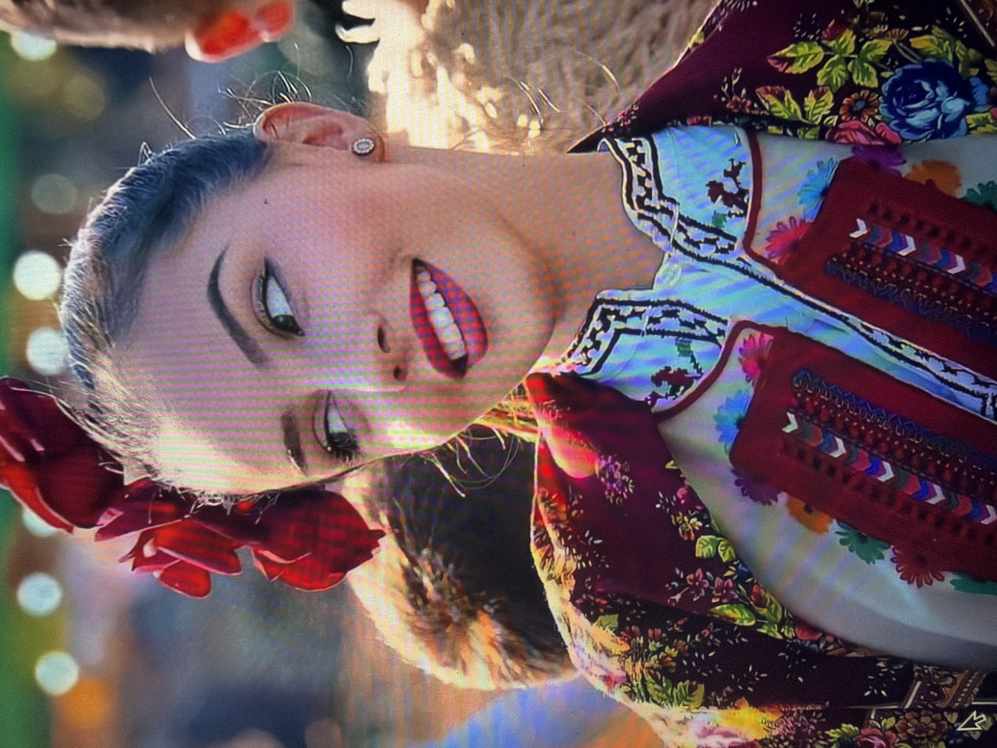 A young female, appearing to be in her late teens or early twenties, is depicted from the upper chest upwards, turned slightly to her left, looking towards her left with a wide smile revealing white teeth. Her dark hair is pulled back into an updo, adorned on the left side of her head with multiple red floral elements resembling roses. She has dark, defined eyebrows, dark eyeliner, and vibrant red lipstick. A small, round, light-colored stud earring is visible in her right ear. She is dressed in traditional Bulgarian attire, featuring a light-colored inner garment with intricate black and white geometric embroidery around the collar and a red chest panel adorned with colorful patterns and white chevron accents. Over this, she wears a dark maroon outer garment, possibly a jacket or shawl, patterned with vibrant green leaves and various colored flowers including blue, pink, and yellow. The background is significantly blurred, displaying out-of-focus lights in green, orange, and light blue hues, indicative of an outdoor or event setting. A portion of another person's head is partially visible to the right in the blurred background. The scene is located in the city of Shumen, Bulgaria.
