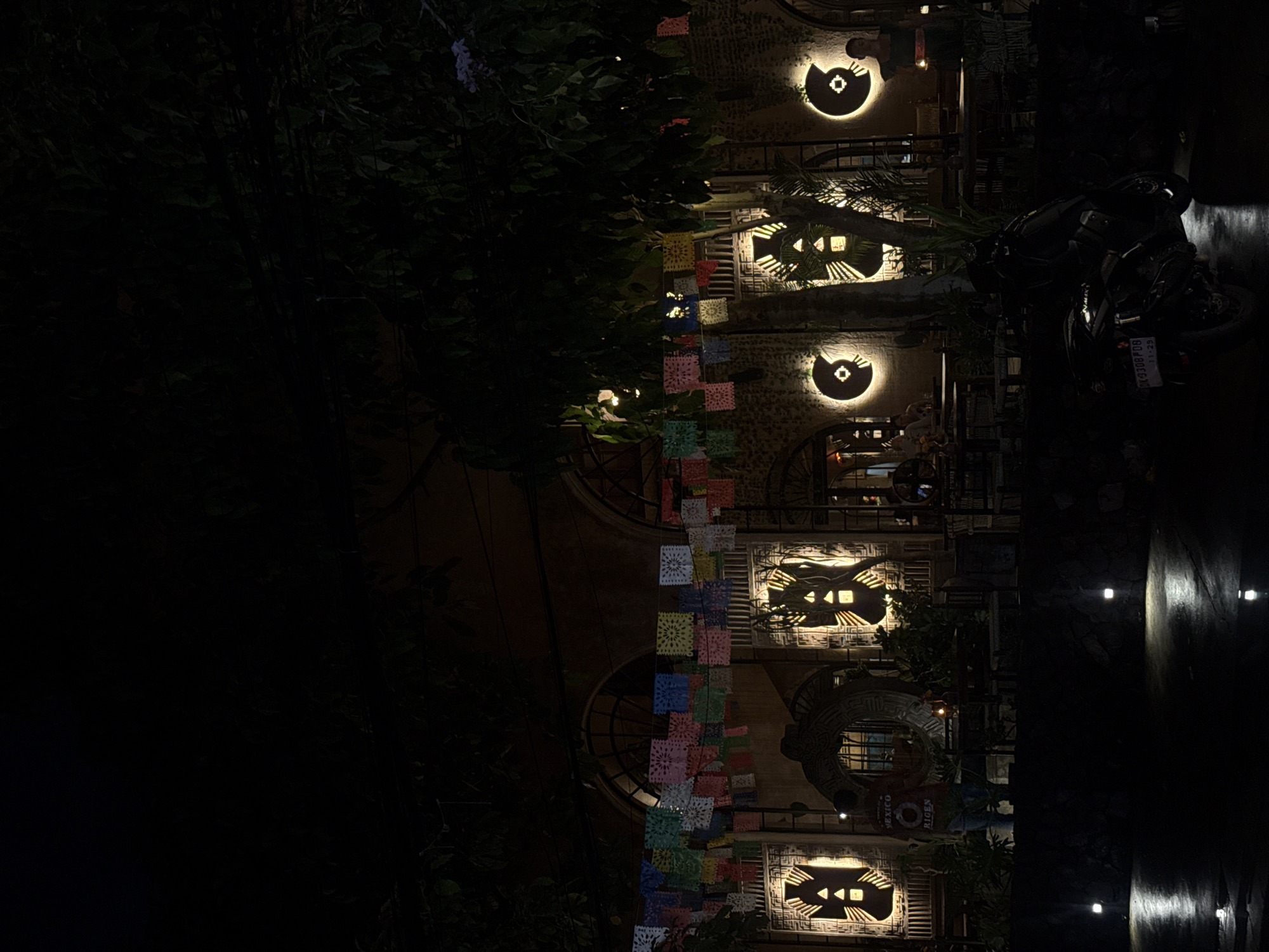 Captured in Unknown, Unknown at night, the image depicts a multi-story building adorned with decorative lights and colorful papel picado banners. The architecture features arched windows and what appears to be stone or textured walls. A motorcycle is parked nearby. Some people are visible in the background. Foliage partially obscures the building. Street lights are visible.