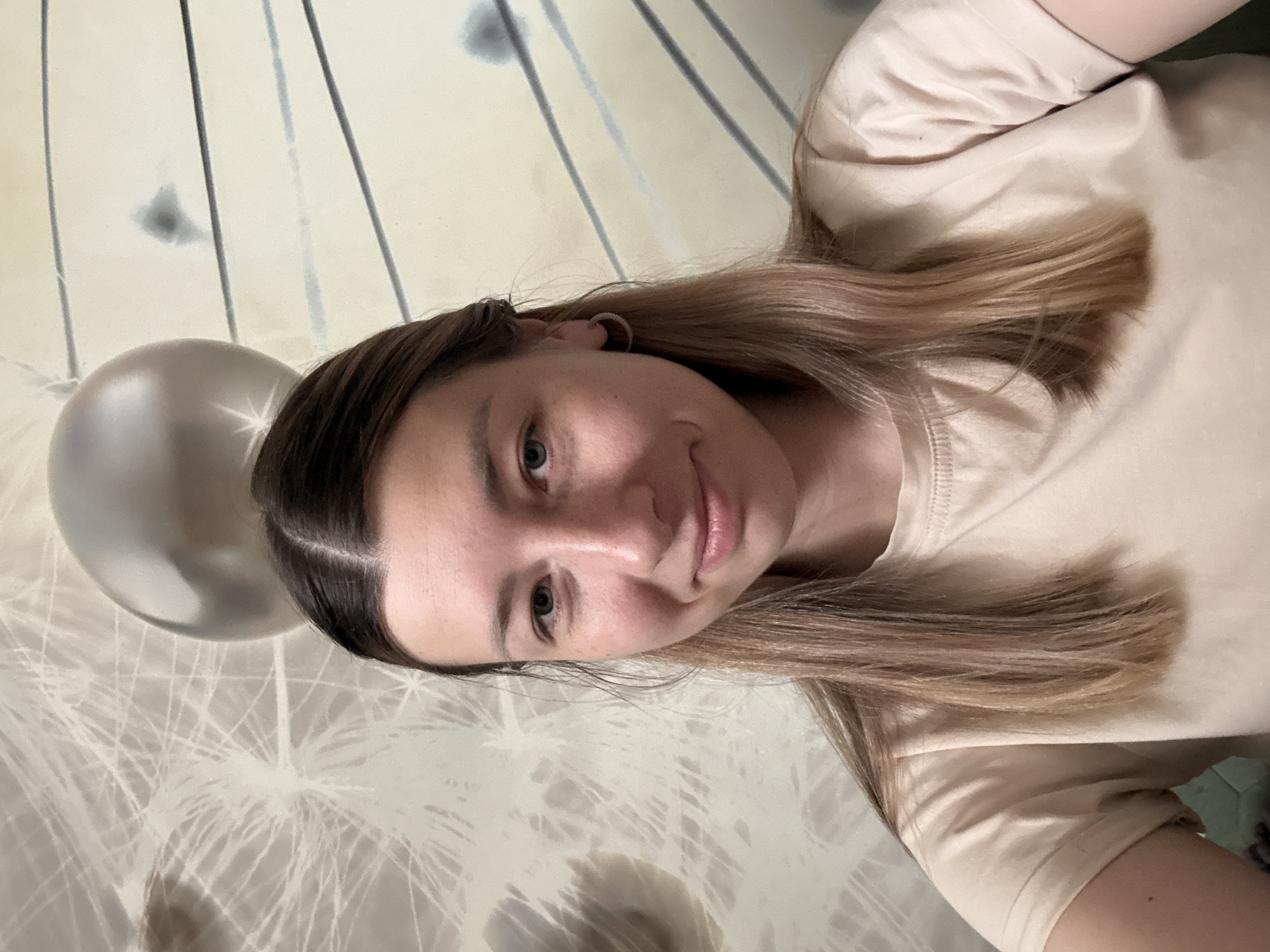 A young woman is featured in a close-up, angled selfie, appearing to be lying down or leaning back. She has light brown hair parted down the middle and blue-green eyes, looking directly at the camera with a soft, subtle smile. She wears a plain, light beige t-shirt and a small hoop earring in her visible ear.
In the upper left corner of the background, a silver-colored balloon is partially visible, showing a bright glint from reflected light. The wall behind her is light-colored and features an abstract pattern of thin, dark lines and lighter, feathery or dandelion-like designs. The lighting in the image is soft and even, suggesting an indoor setting. There is no visible text in the image.