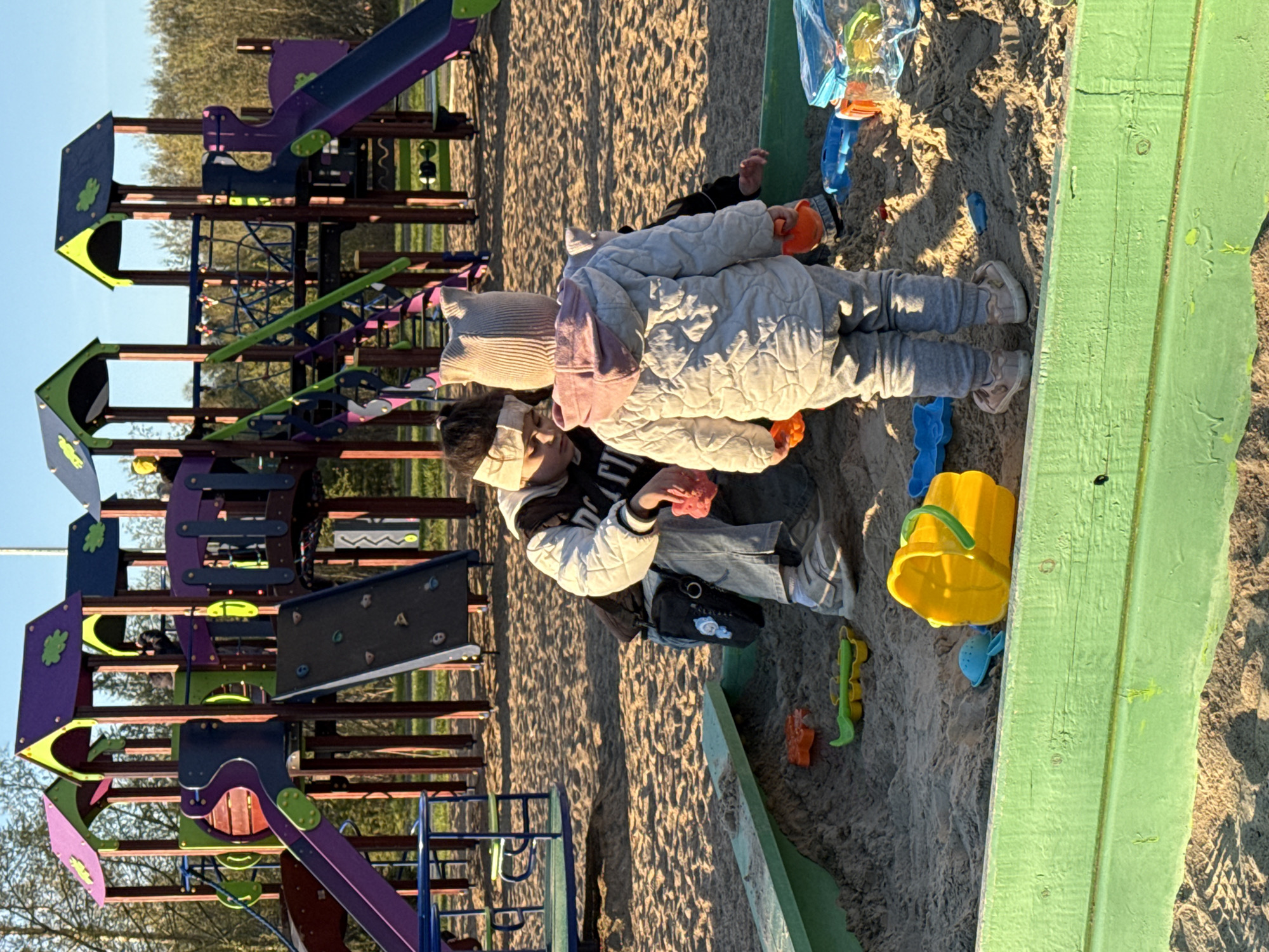 A photograph taken in Tobolsk, Russia, shows two children playing in a sandbox next to a playground. The playground structure is multi-colored, featuring slides, climbing areas, and platforms. One child, presumably an older sibling, is kneeling next to the sandbox and wearing a white jacket, jeans, and a black bag. The second child, possibly a younger sibling, is wearing a tan quilted jacket, grey pants, and a tan knit hat with a pink stripe. Various plastic toys, including buckets, shovels, and molds, are scattered around the sandbox. The sandbox has a green painted wooden frame. The environment suggests a public park on a sunny day.