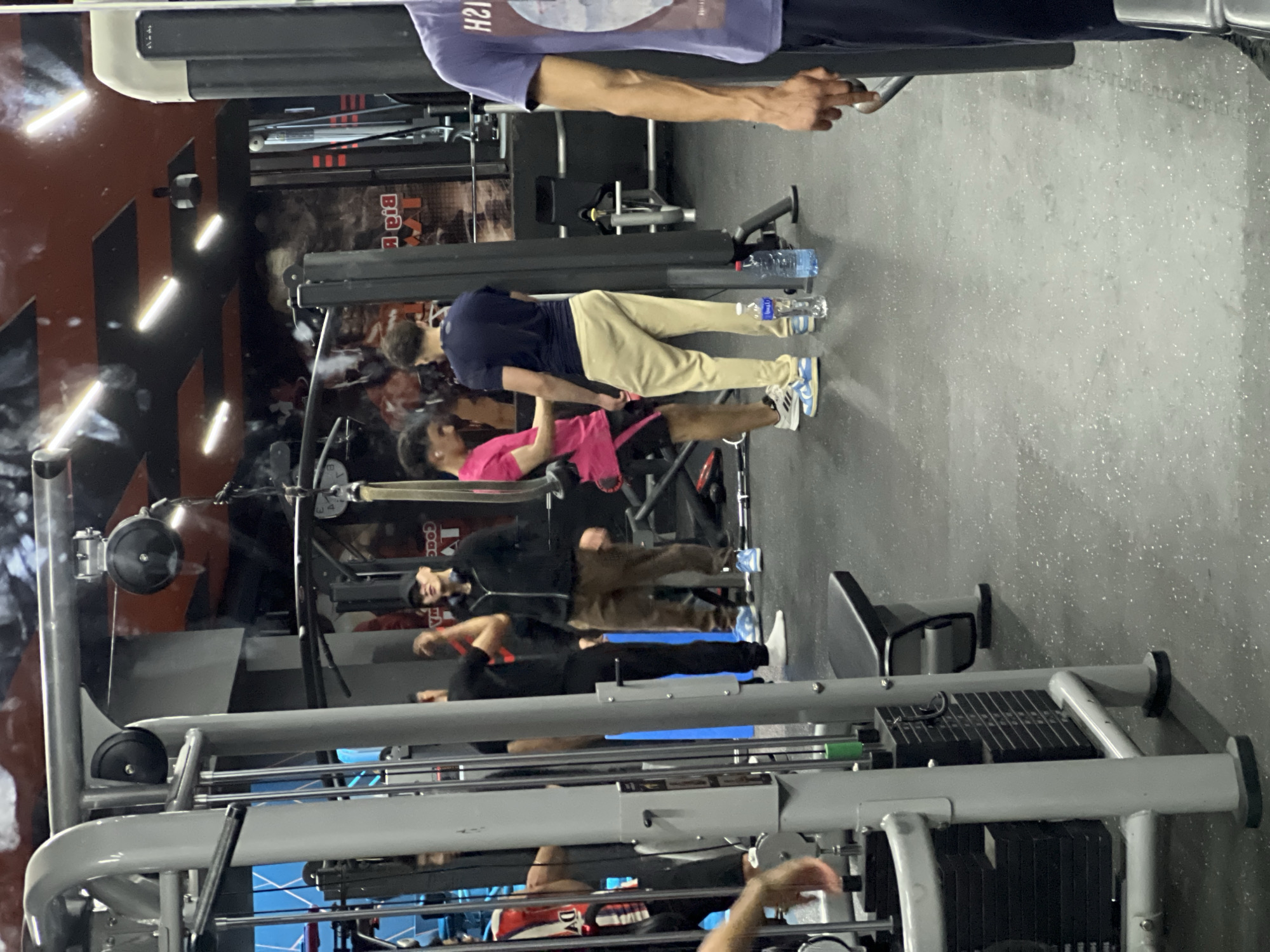The image shows the interior of a gym. There are several people engaged in various exercises on different gym equipment. The gym has weights, cable machines, and other fitness apparatus. The background includes walls with decorative elements and lighting fixtures. Without further identifying information or landmarks, it is not possible to confirm the location within M'Diq.
