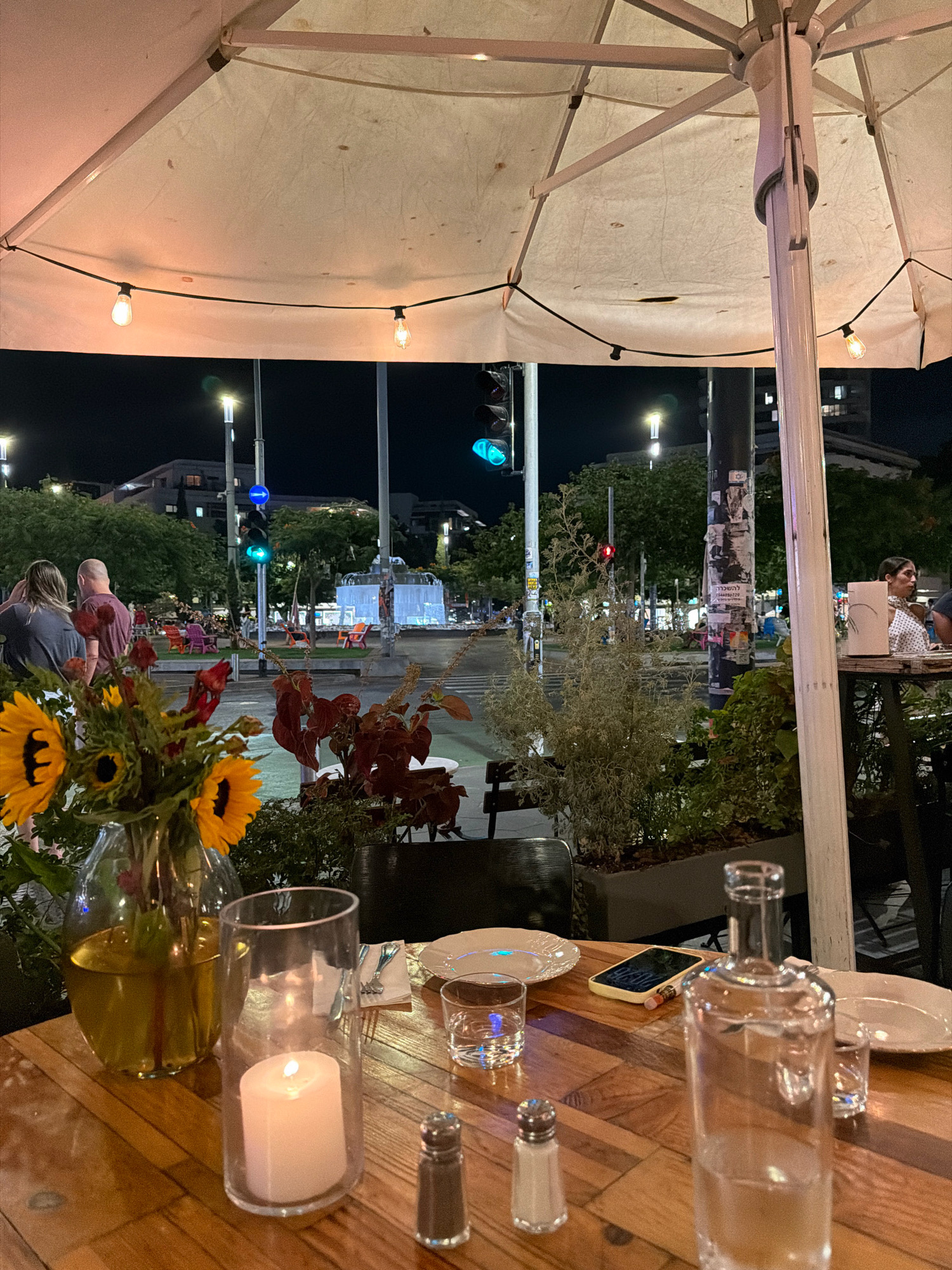 The image depicts an outdoor dining scene at dusk in Tel Aviv, Israel, likely near Rothschild Boulevard, given the traffic lights and urban setting. The foreground is dominated by a wooden table laden with dining essentials: a glass vase holding sunflowers and other flowers, a white pillar candle in a glass holder, salt and pepper shakers, a smartphone, plates, and a glass bottle of water. Beyond the table, a large white umbrella provides overhead cover, with string lights casting a warm glow. In the background, a street scene unfolds with traffic lights showing green and red signals, pedestrians walking, and colorful chairs arranged in a public space, possibly a park or plaza. Buildings and trees line the street, illuminated by ambient light.