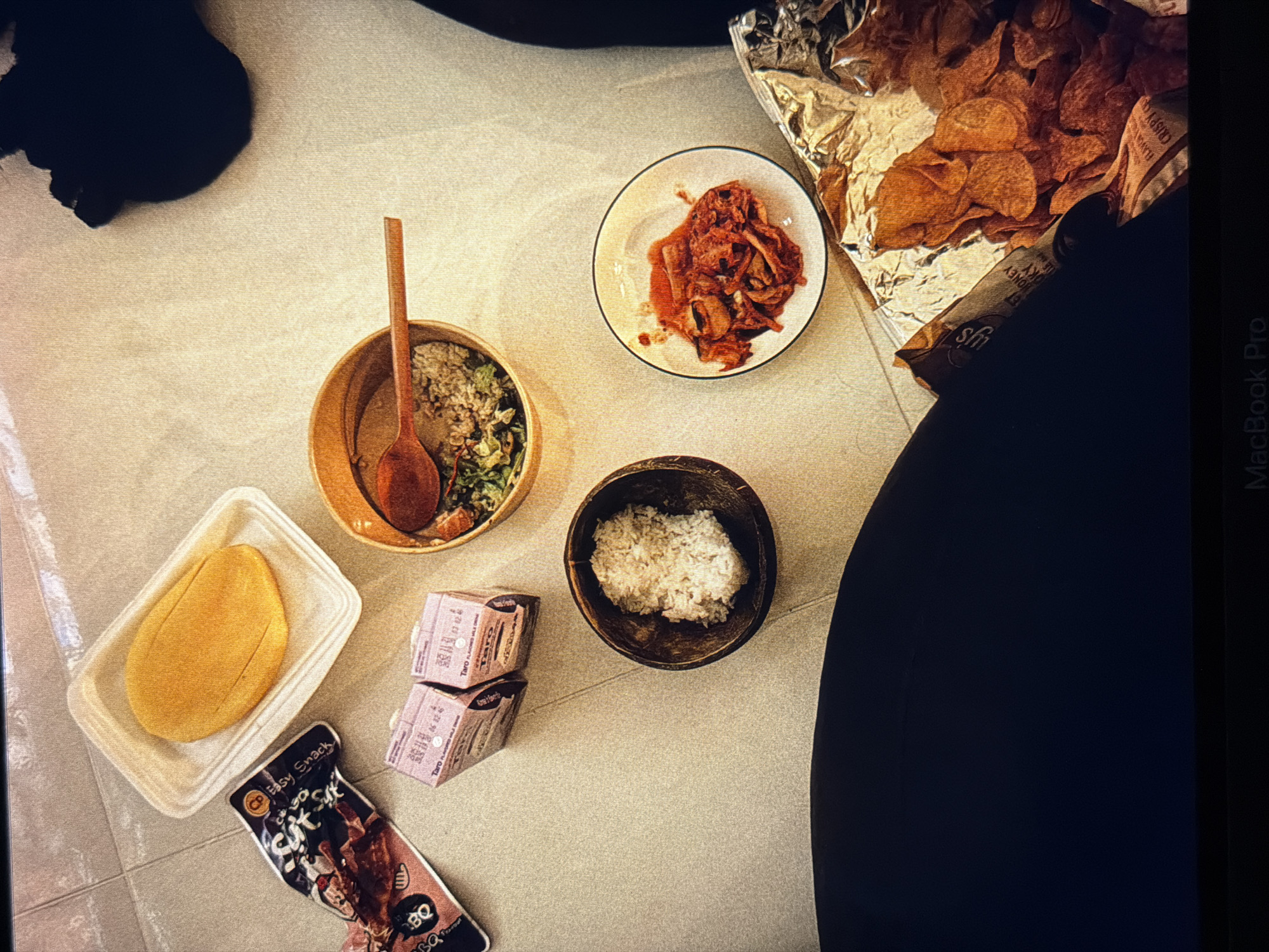 The image depicts a ground-level food arrangement. There is a bowl of rice, a bowl of salad with a wooden spoon, a small plate of kimchi, and an opened bag of Lay's potato chips. Two small cartons of Taro-flavored milk, a mango in a plastic container, and a CP Easy Snack (BBQ flavor) packet are also visible. Parts of a person in dark clothing (possibly legs) are visible on the left and right edges of the frame. A black object is in the upper left, possibly a pet. "MacBook Pro" is printed in small letters on the right edge. The scene appears to be indoors, on a light-colored, tiled floor. No specific location in Đà Nẵng, Vietnam, is definitively indicated.