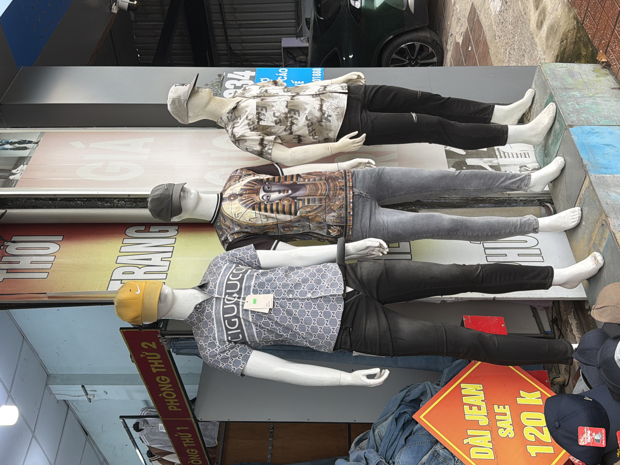 The image displays a storefront in Nha Trang, Vietnam, featuring three male mannequins dressed in casual attire. The mannequins are positioned in a line, with the leftmost wearing a patterned short-sleeved shirt and dark jeans, the middle mannequin in a graphic t-shirt featuring an Egyptian pharaoh and grey jeans, and the rightmost mannequin in a patterned short-sleeved shirt and black ripped jeans. All mannequins are wearing baseball caps.
In the foreground, several baseball caps are displayed on a surface. To the left of the mannequins, a bright orange sale sign with Vietnamese text is visible, indicating "DAI JEAN SALE 120 K". The background shows elements of a shop interior, including clothing racks with garments, and signage with Vietnamese text. A portion of a dark-colored car is visible on the right side of the image. The overall scene suggests a retail environment focused on men's fashion.