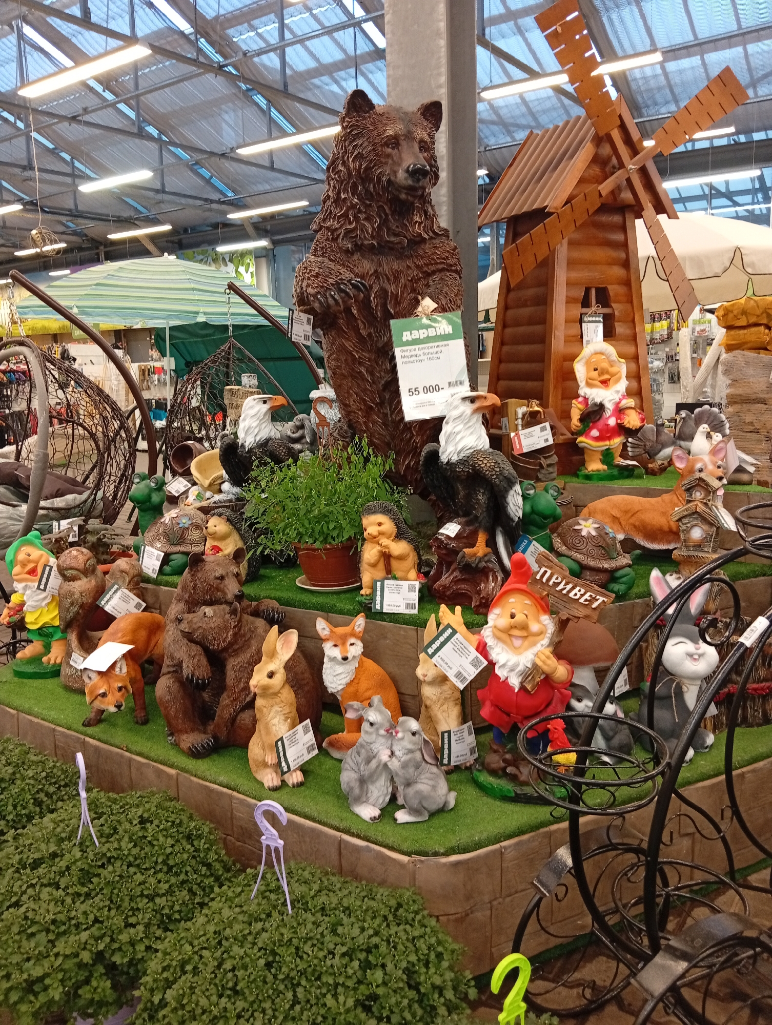 An indoor retail environment, likely a garden center or home goods store, displays a collection of garden ornaments. The scene is dominated by numerous decorative figurines crafted to resemble animals and fantasy creatures, arranged on tiered platforms covered with artificial grass. Prominent among the displays is a large, detailed sculpture of a brown bear standing upright, with a price tag visible. To its right, a rustic wooden windmill structure is present. Other figurines include gnomes, eagles, foxes, rabbits, a turtle, a hedgehog, a frog, and a corgi dog. Some figurines are holding or interacting with smaller objects like birdhouses. The background reveals shelving units with various products, industrial lighting fixtures on the ceiling, and large green and striped umbrellas, suggesting an indoor market setting. The overall impression is one of a commercially organized display of decorative items. No people are visible in the image. The image was taken on an unspecified date in Pushkino, Russia.