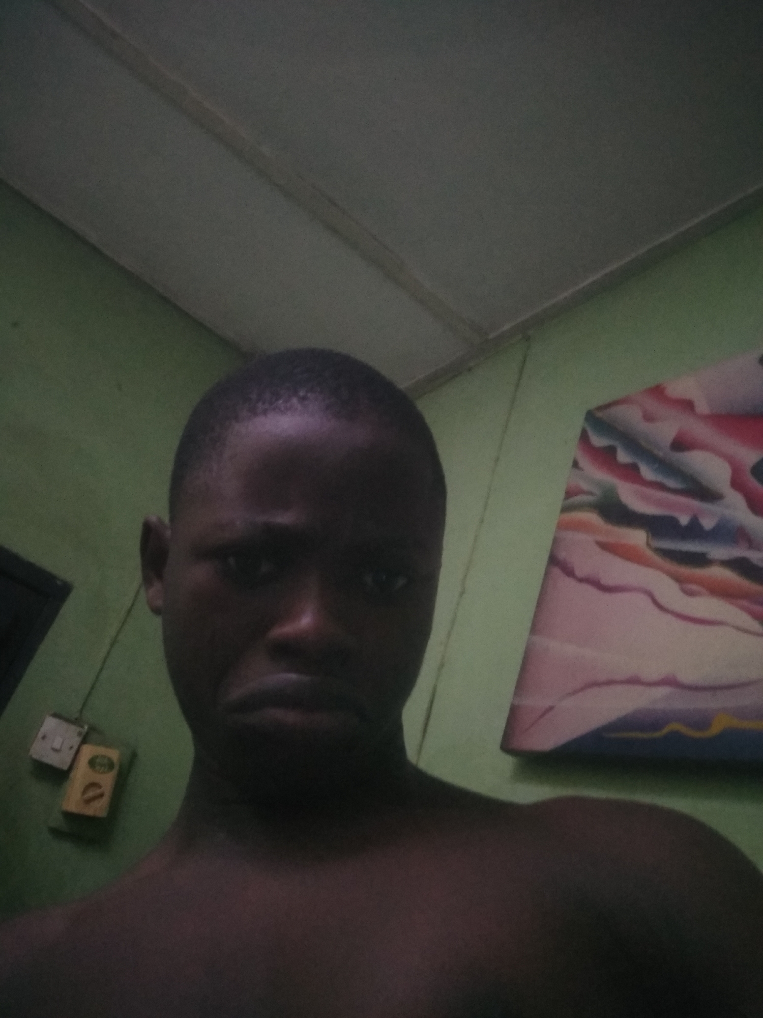 A portrait-style image captures a young Black man from the chest up, positioned indoors. He appears to be in his late teens or early twenties, with short dark hair. His skin tone is dark, and he is either shirtless or wearing a dark top that blends with his complexion in the dim lighting. His facial expression is somber, featuring a slight frown and downcast eyes, suggesting sadness or solemnity.
The background includes a light green painted wall and a dark gray ceiling. On the left side of the wall, there is a white light switch and a small, rectangular yellow box, possibly an electrical component. Faint, unreadable text or symbols, resembling "01" or "OD", are partially visible on the yellow box. To the right, a portion of a colorful abstract artwork or painting is visible, displaying swirling patterns in shades of pink, blue, orange, and white.
The lighting in the room is dim and artificial, creating a moody atmosphere. The setting appears to be a residential indoor space. No specific activity is taking place other than the man posing for the camera. The context suggests the location is Shangisha, Nigeria.