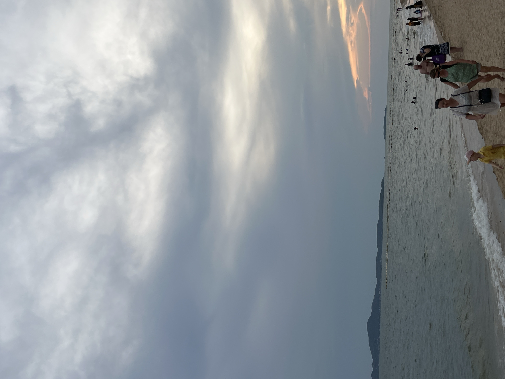 The image shows a beach in Pattaya City, Thailand. The sky is overcast with some bright areas. A line of people is walking and standing on the beach. The water is calm with small waves breaking on the shore. A distant coastline is visible in the background.