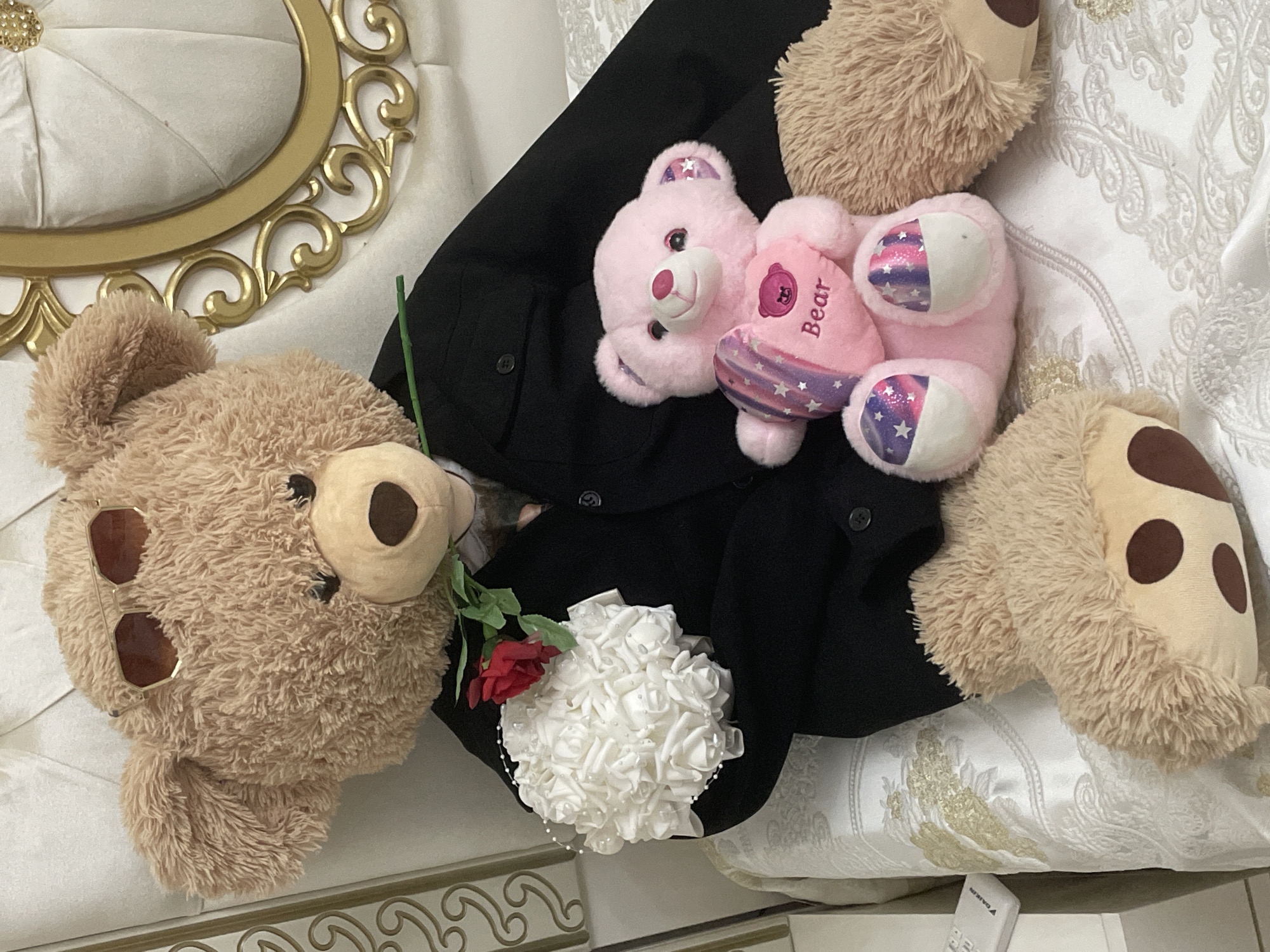 An overhead shot displays a collection of items arranged on white and gold ornate furniture, likely indoors in Ihtiman, Bulgaria.
The central focus is a black garment, possibly a jacket or blazer, draped horizontally. On top of this garment, to the right, lies a small, pink teddy bear. This bear has a light pink snout and ears with a starry, iridescent purple-blue fabric peeking through. It holds a pink heart-shaped pillow embroidered with the word "Bear" in darker pink. Its paws also feature the same starry, iridescent fabric pattern.
To the left of the black garment and partly overlapping it, a larger, light brown teddy bear with shaggy fur is positioned. This bear wears a pair of gold-rimmed sunglasses on its head and has a light brown snout and dark brown patches on its face, resembling eye patches.
Between the two bears, on the black garment, there is a small, artificial bouquet composed of numerous white roses, possibly with small pearl accents. A single red rose with a green stem is placed beside this white bouquet, leaning against the larger brown bear.
The background consists of richly upholstered furniture, featuring white fabric with intricate gold patterns, suggesting a classic or ornate style. Part of a white object, identified as a "DAIKIN" remote control, is partially visible in the bottom right corner. The lighting suggests an indoor setting with no specific time of day indicated. The arrangement appears staged, possibly for a decorative or gift-giving purpose.