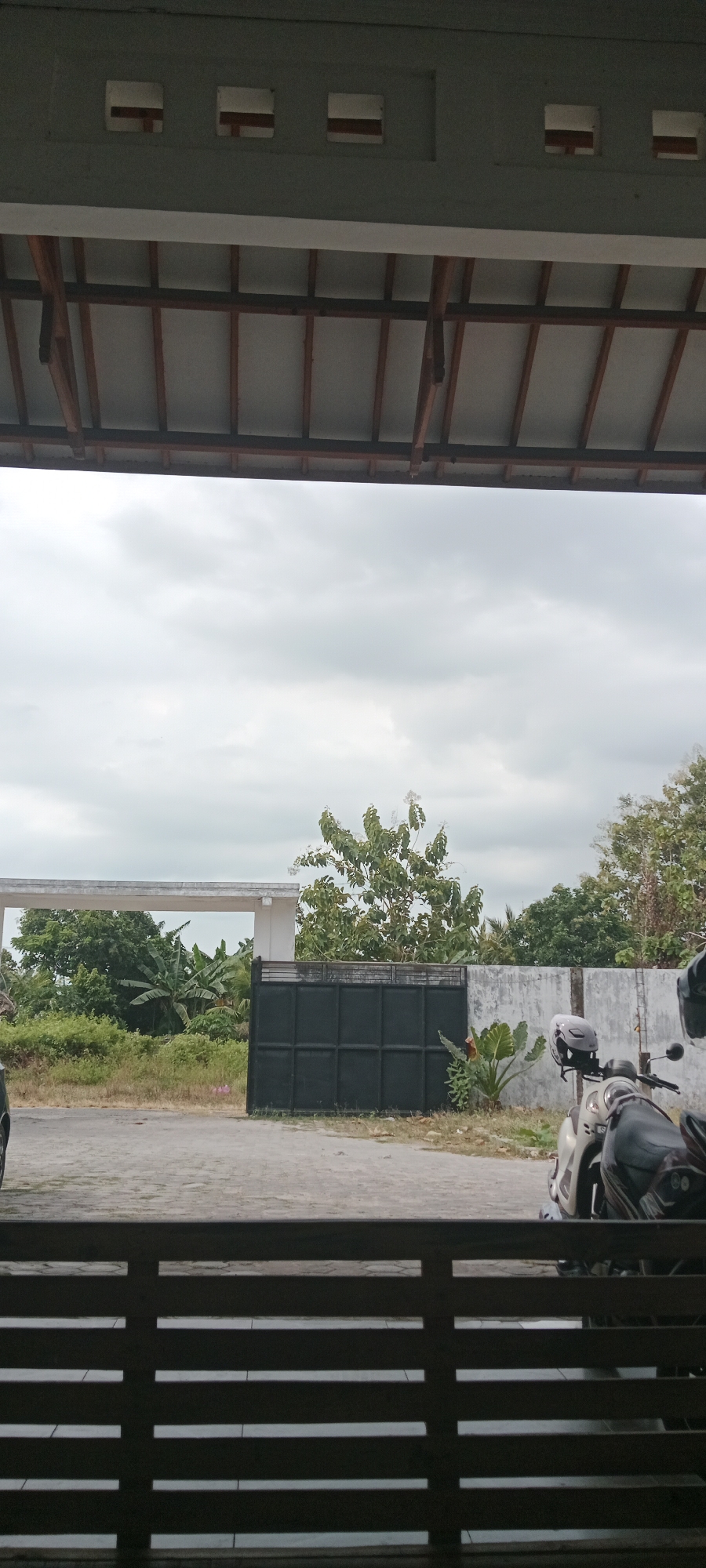 The image depicts a cloudy sky above a residential area in Tuban, Indonesia. In the foreground, a dark wooden railing with horizontal slats is visible, suggesting a porch or balcony. Beyond the railing, a paved driveway leads to a black metal gate. To the left of the gate, a white building with a flat roof is partially visible, surrounded by lush green vegetation, including banana trees and other tropical plants. To the right of the gate, a concrete wall with a textured surface runs along the property. A motorcycle, with a helmet resting on its seat, is parked next to the wall. The overall scene conveys a sense of a typical Indonesian residential environment under an overcast sky.