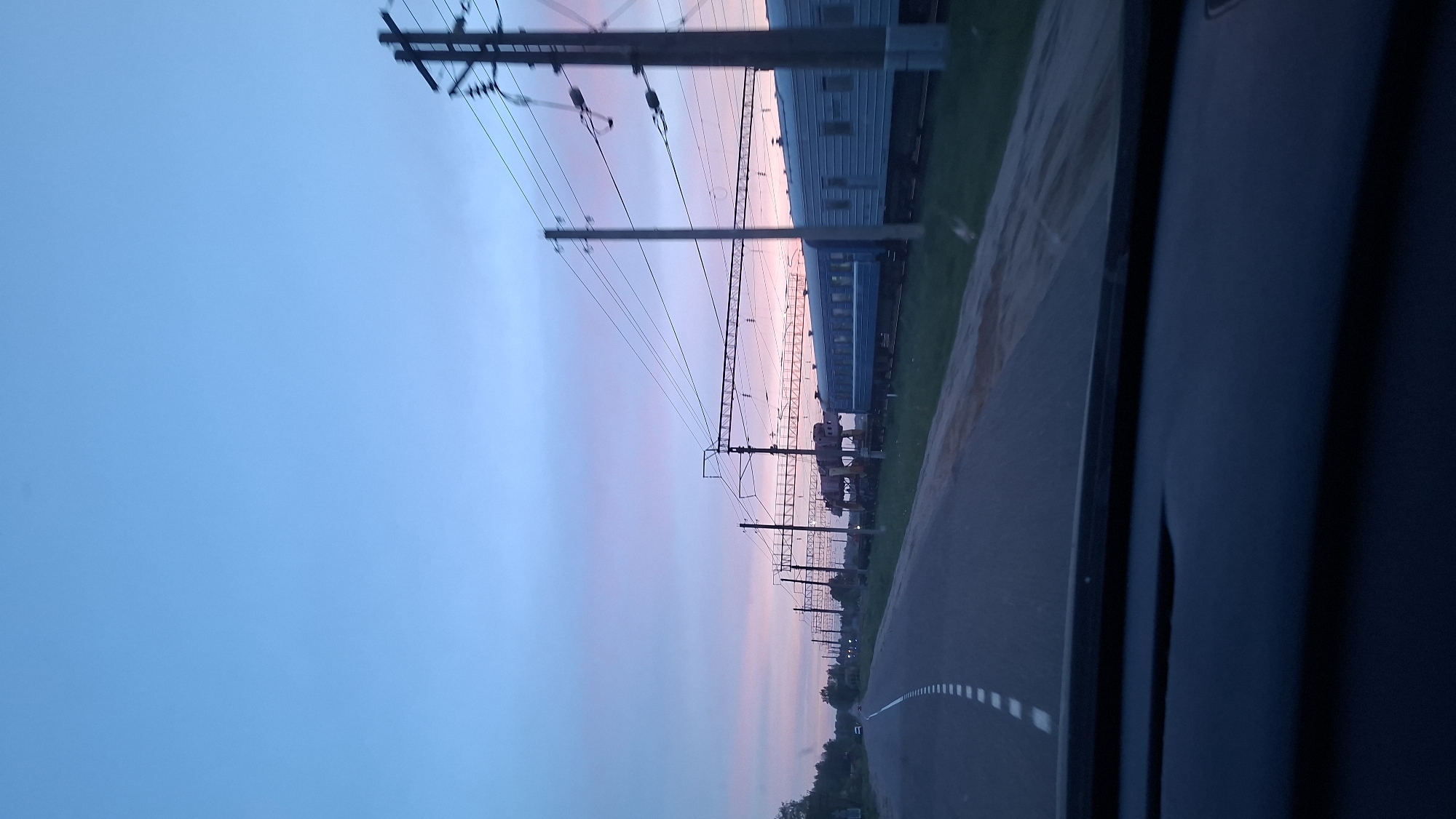 Captured from a high angle, the image presents a landscape view from a moving vehicle. The setting is a street lined with utility poles, their wires stretching across a serene sky, tinged with hues of pink and blue, indicative of either dawn or dusk. Adjacent to the road is a railway track where a blue train car is visible. The image, likely taken in Unknown, Unknown, is partially obstructed by the vehicle's interior frame in the foreground, creating a sense of movement.