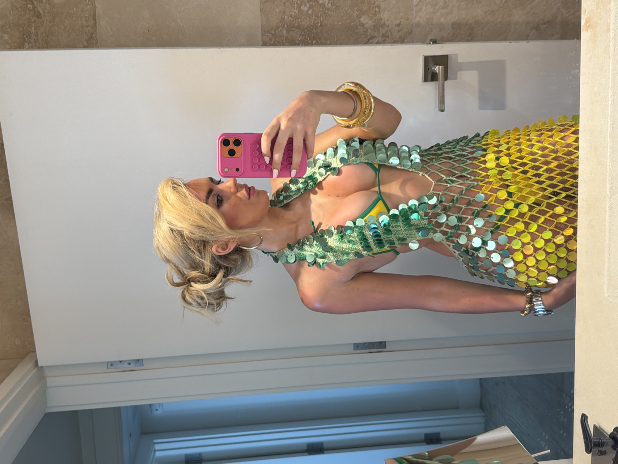 A woman is taking a selfie in a bathroom. She is wearing a green and yellow sequined dress and gold jewelry. She is holding a pink smartphone in her right hand. The bathroom has white walls and a light-colored tile floor. The bathroom includes a chrome faucet. The location is Miami Beach, United States of America.