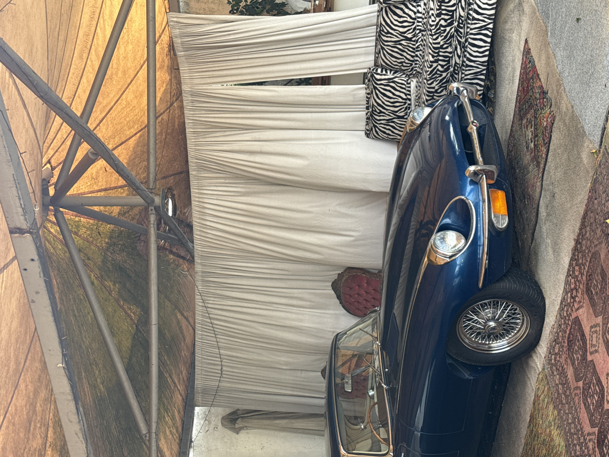 The image shows a blue vintage car parked indoors under a tent-like structure, possibly a garage or event space, in Kraaifontein, South Africa. The tent has a metal frame with a tan fabric roof. There is white fabric draped as a backdrop behind the car, and zebra-patterned cushions and a red chair are visible. A patterned rug sits adjacent to the car. The scene suggests a possible display or event setting for the vintage car.