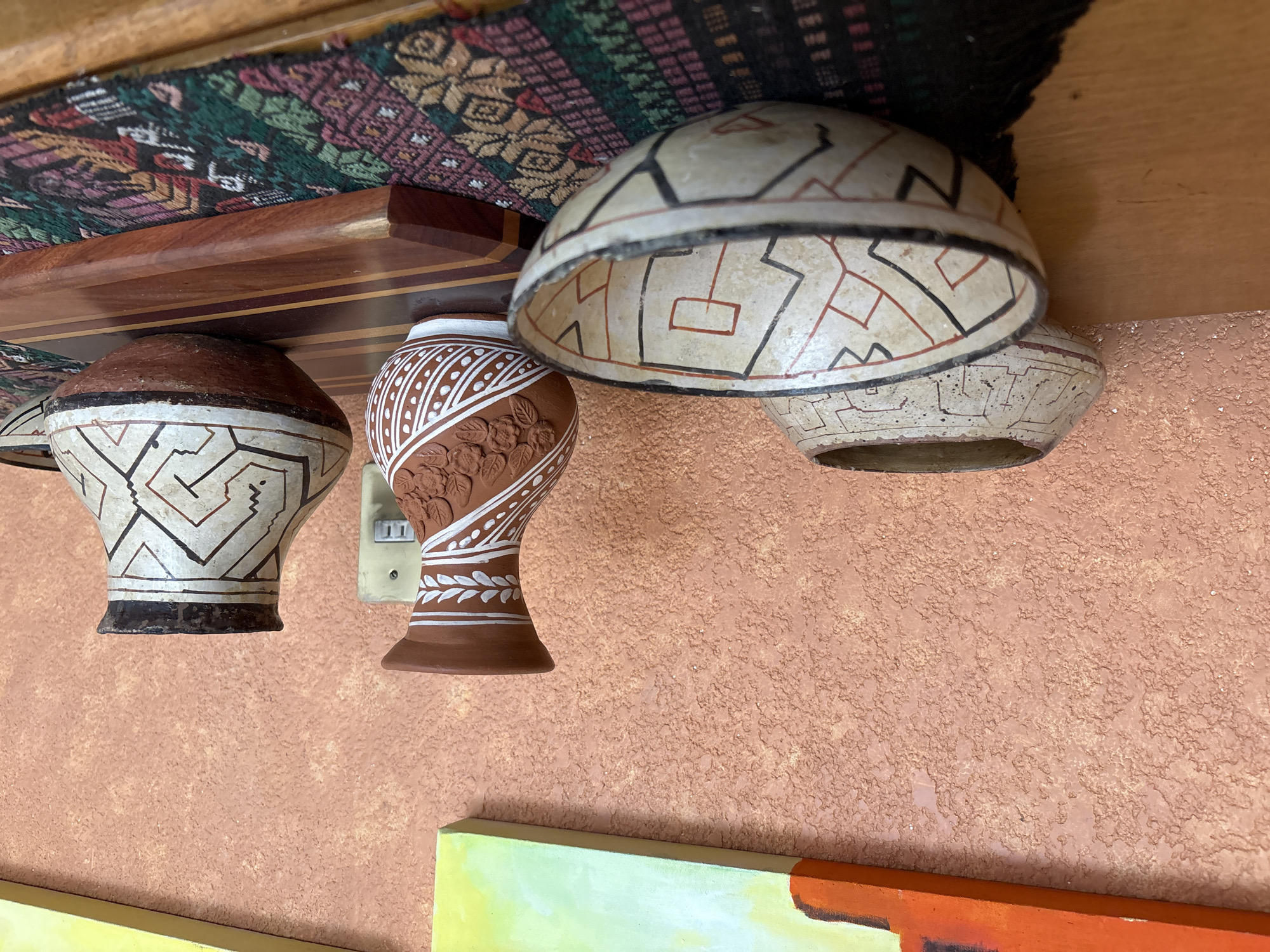 The image shows a set of four vases or pots, suspended from a wooden shelf that has a patterned textile lining the back. The pots vary in design, with some showcasing geometric patterns and others floral. The wall behind them is textured and painted in an orange hue. In the lower part of the image, there's a glimpse of artwork with areas of yellow and orange. There are no identifiable people or specific actions taking place. The location is Unknown, Unknown.