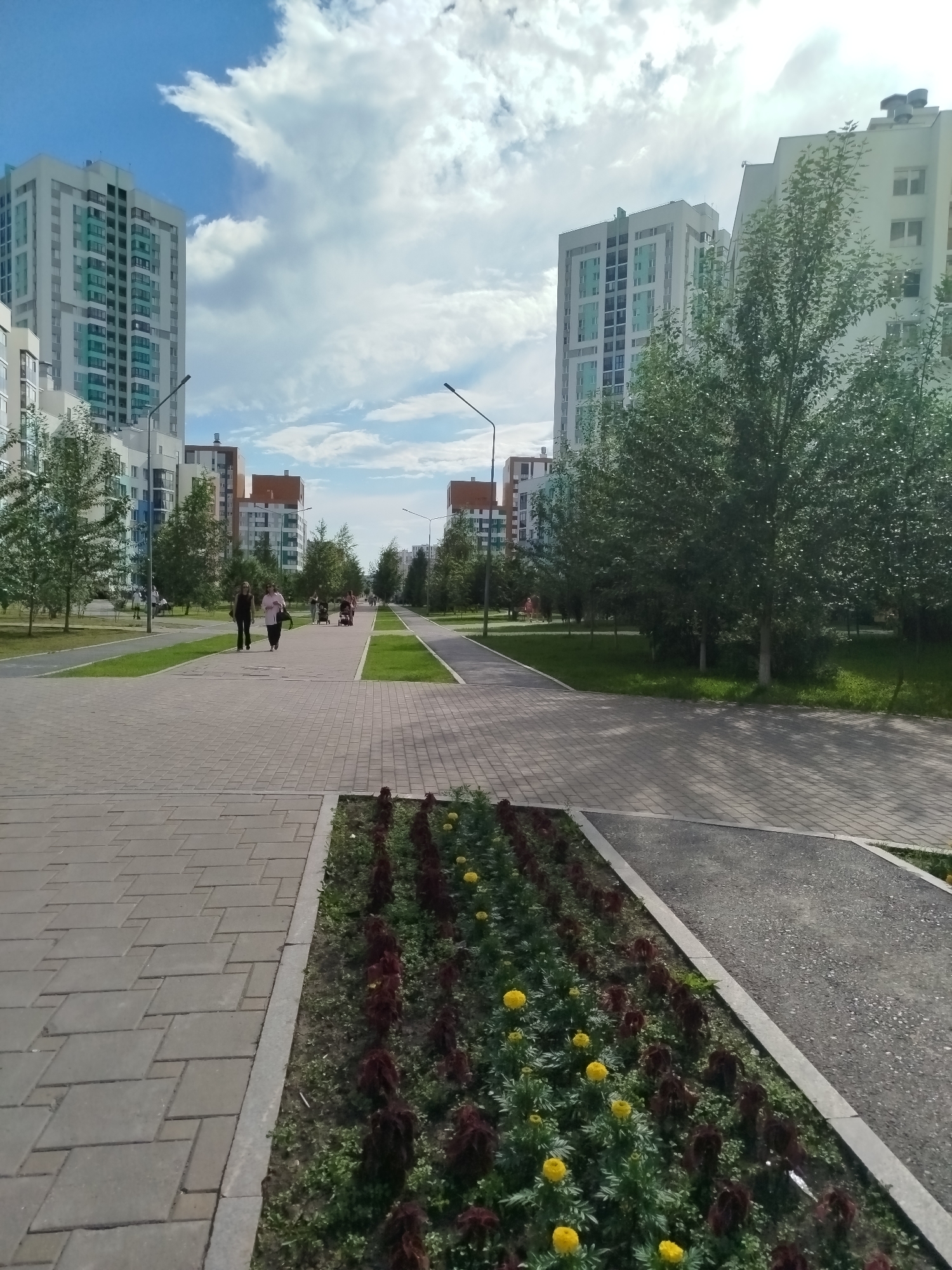 The image depicts a modern urban park in Yekaterinburg, Russia, on a bright, partly cloudy day. In the foreground, a paved walkway with a textured surface made of light gray interlocking stones leads into the distance. To the right of the walkway, a flower bed runs alongside it, featuring rows of dark red plants and vibrant yellow marigolds.
Beyond the flower bed, a paved path for a different purpose, possibly a bicycle lane or a secondary walkway, is visible, separated by a strip of green grass. The background is dominated by tall, modern apartment buildings with light-colored facades and many windows. Trees with lush green foliage are planted along the walkways and in grassy areas between the buildings, suggesting a well-maintained urban green space.
Several people are strolling on the main walkway, including two women walking together, one in a dark outfit and the other in a light pink top. Further in the distance, other individuals and what appear to be strollers or small carts suggest a family-friendly environment. Streetlights are also visible, indicating the park is equipped for evening use. The clear blue sky with scattered white clouds and the sun casting shadows on the ground suggest it is daytime, likely late morning or early afternoon. The overall impression is one of a peaceful and well-developed residential area with ample recreational space. No visible text is present in the image.