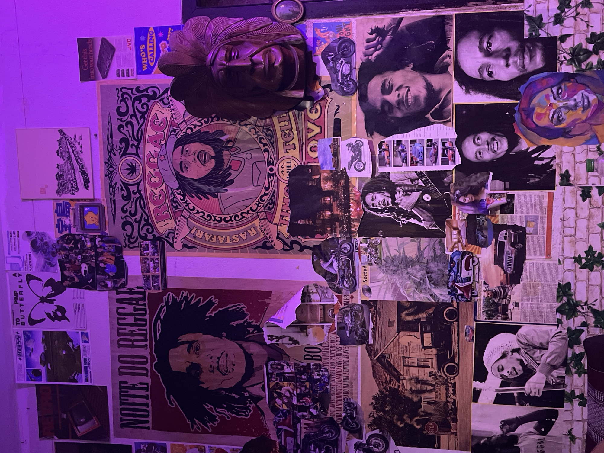 This image shows a wall covered in a collage of posters and artwork. The dominant subject matter is Bob Marley, with multiple portraits of the reggae artist displayed. Other images include a large wooden mask with a headdress, motorcycles, cars, houses, and a marijuana plant. Some text on the posters is in Portuguese, indicating "Noite do Reggae" (Reggae Night). The lighting is purple, suggesting ambient club or room lighting. The wall appears to be brick. The presence of a "Noite do Reggae" poster suggests a connection to reggae music culture. The city of Chalong in Thailand is known for its vibrant nightlife and bars, some of which might feature such decor to create a specific ambiance.