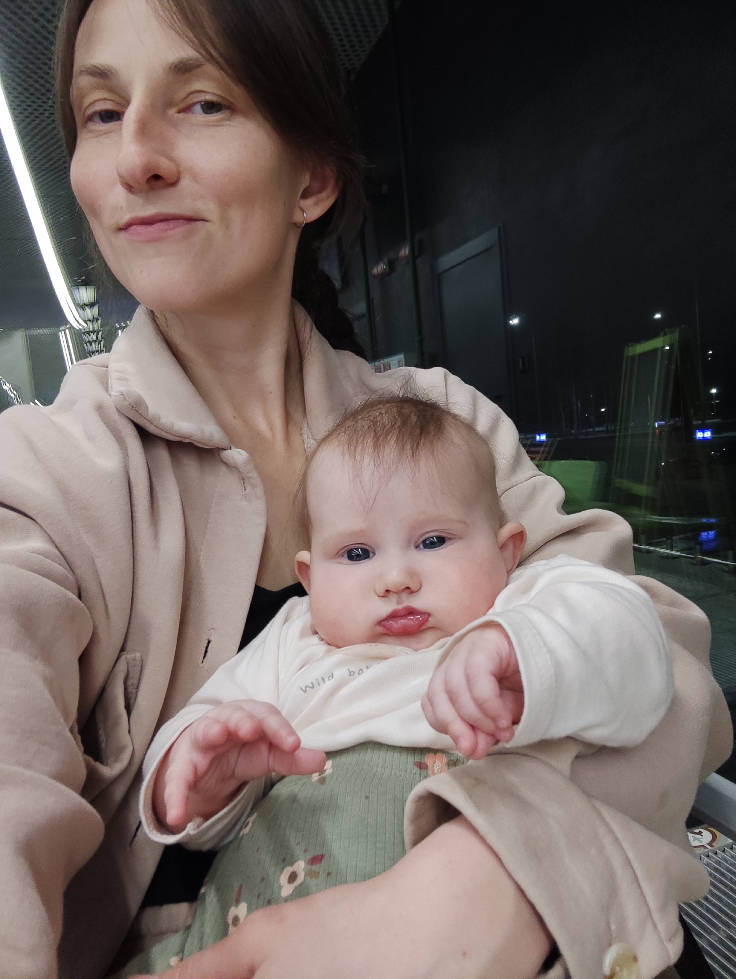 An adult female is depicted holding an infant in what appears to be a selfie, taken indoors at night. The setting is identified as the Izhevsk Railway Station (Izhevsk Passazhirsky) in Izhevsk, Russia, based on distinctive architectural elements visible through the background window.
The female subject, likely in her late 20s to early 30s, possesses light skin and brown hair pulled back, with a visible braid on her right side. She wears a small silver hoop earring in her left ear. Her attire consists of a light beige collared jacket over a dark undershirt. She maintains direct eye contact with the camera, exhibiting a slight, closed-mouth smile.
The infant, estimated to be between three and six months old, has light skin, sparse light brown hair, and blue eyes. It wears an off-white long-sleeved top with partial text "Wild bo..." visible on the chest, and green ribbed overalls adorned with a faint floral pattern. The infant's mouth is slightly pouted, and it also looks directly at the camera, held securely by the woman's left arm. The infant's right hand is slightly open, fingers curled.
The immediate indoor environment includes a metallic, possibly slatted or perforated ceiling structure with linear lighting fixtures visible above the subjects. A grey metal grille, resembling part of a seat or barrier, is partially visible to the right of the woman.
Through a large glass partition or window behind the subjects, a dark exterior is visible, indicating nighttime. Distant lights are scattered across the dark background. A prominent green, skeletal-like structure, possibly scaffolding or an architectural feature, is discernible on the right side of the outdoor view. Reflections on the glass reveal faint blue signs with white Cyrillic text, consistent with a public transport facility.