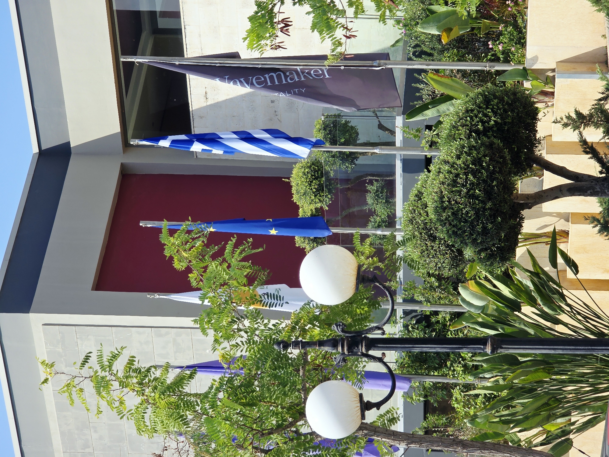 The image shows an exterior view of a building with a modern design, featuring a grey concrete facade and a prominent maroon-colored section. Several flags are displayed in front of the building. A Greek flag with blue and white stripes is prominently visible. To its left, a dark grey flag with white lettering, partially obscured, reads "Govemaker" and "Hospitality". To the left of the Greek flag, a blue flag with yellow stars, likely the European Union flag, is also displayed. Further to the left, another flag with a green branch on a white background is visible. The building is surrounded by lush greenery, including trees with dense foliage, manicured bushes, and large-leafed tropical plants in planter boxes. A black lamppost with two white globe lights stands in the foreground, adding to the scene's aesthetic. The overall environment suggests a well-maintained public space or the entrance to a hospitality establishment in Daratsos, Greece. There are no people visible in the image.