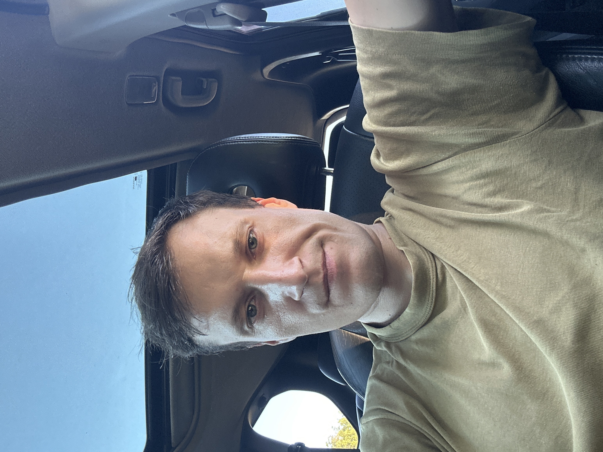 A man in a tan t-shirt is sitting in the driver's seat of a car. The car has a sunroof that is open, and the sky is visible through it. The man is looking at the camera and smiling. The interior of the car is dark, but some of the dashboard and seats are visible. The man's hair is dark and he has blue eyes. The image appears to be taken during the daytime. The location in the city of Makiv, Ukraine is not specified in the image.