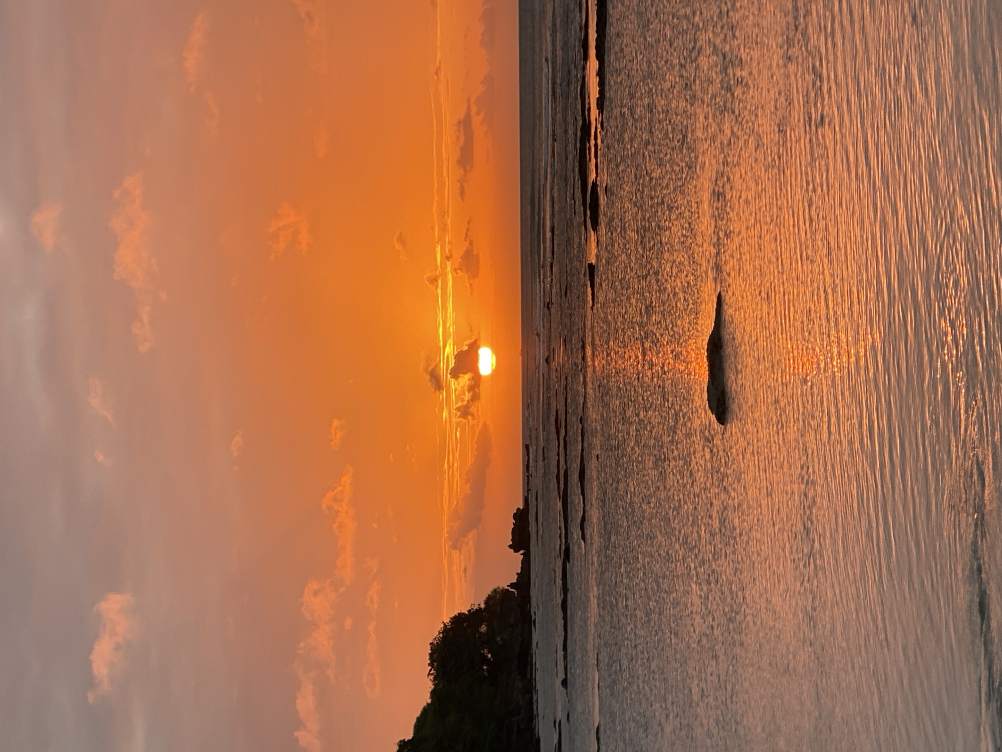 A wide shot captures a sunset over the ocean with a serene, orange glow filling the sky. The sun is partially obscured by clouds, casting a shimmering reflection on the water. The foreground features calm water with subtle ripples, and a lone dark rock sits in the middle. In the background, a rocky shoreline with lush green trees is visible on the left. The scene appears to be a peaceful coastal landscape in Kuta Selatan, Indonesia.