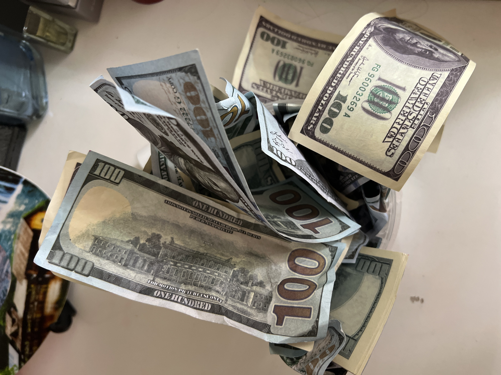 The image depicts a clear glass jar filled with numerous US $100 bills. Some of the bills are protruding from the jar. Visible details on the bills include the portrait of Benjamin Franklin, the phrase "ONE HUNDRED DOLLARS," the Federal Reserve seal, and the serial numbers. The setting appears to be indoors on a white surface, possibly a table or countertop. The lighting is bright. There is no evidence to support that this image was taken in Sochi, Russia.