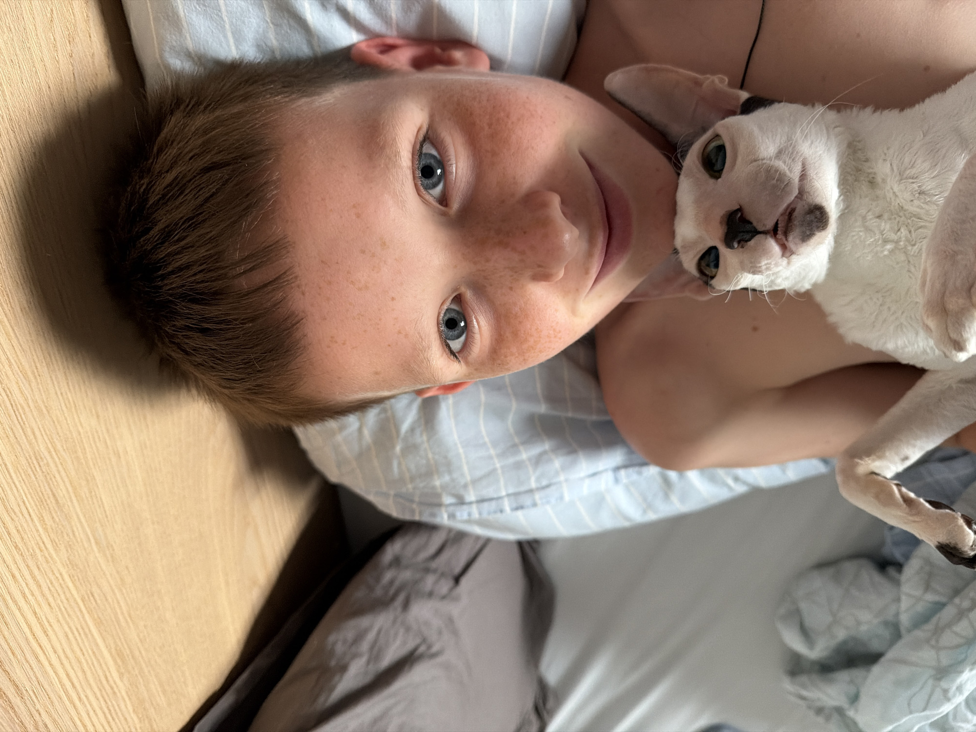 The media file shows a close-up, overhead shot of a young boy and a white cat lying down. The boy has light skin with freckles and blue eyes. His hair is brown and short. He is looking up at the camera with a slight smile. The cat, which is white with black markings on its face and paws, is nestled against the boy, looking towards the camera with its mouth slightly open. The subjects are lying on a bed with blue and white striped bedding. To the left of the bed, a wooden surface, possibly a nightstand, is visible. The lighting suggests it is daytime, likely indoors. The overall mood appears relaxed and affectionate, with the boy and cat appearing comfortable together. There is no visible text in the image. The location context is Yekaterinburg, Russia.