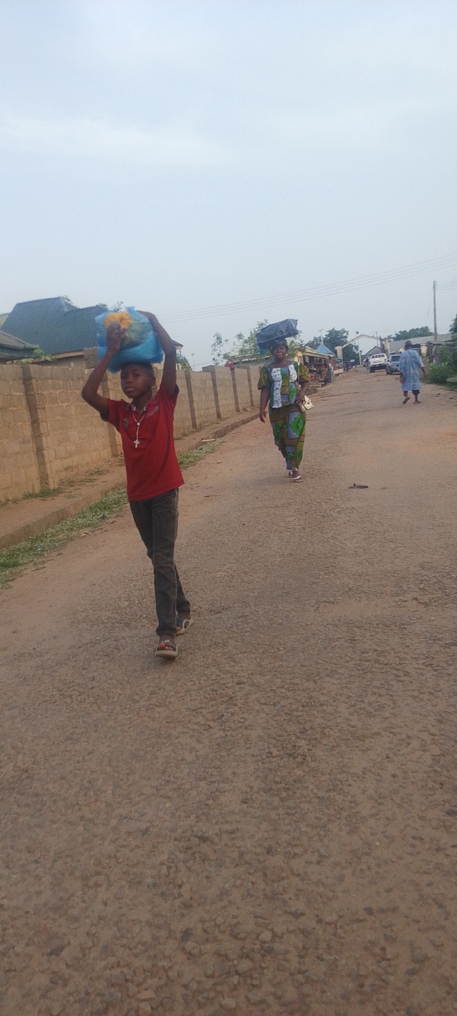 Adankolo, Nigeria: A young boy wearing a red shirt, gray pants, and sandals walks along an asphalt road, carrying a blue bag on his head. He wears a cross necklace. Behind him, a woman in a patterned green and white dress also carries an object on her head. Farther down the road, another person walks away from the camera. In the background, houses, cars, and a building can be seen. A wall runs along the left side of the road. The sky is overcast.