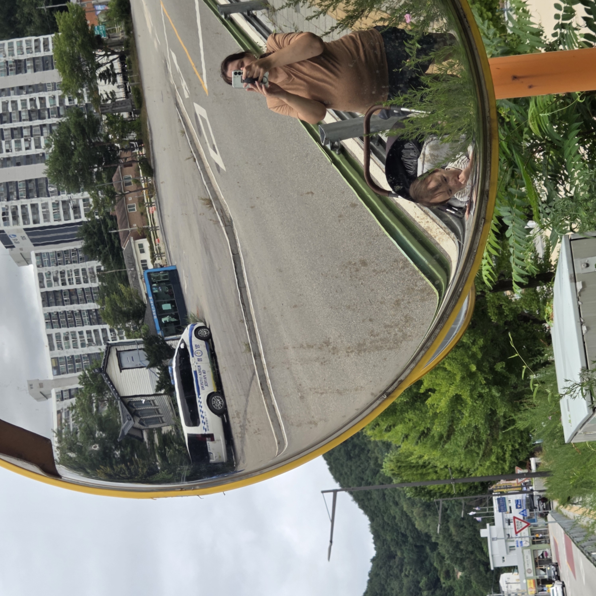 This is a reflection in a convex mirror, likely a traffic safety mirror, situated in Dunnae-myeon, South Korea. The mirror shows a woman holding a smartphone, taking a picture of herself and her surroundings. Beside her, reflected in the mirror, is a baby in a stroller. The background of the reflection displays a street scene with a white police van bearing Korean characters, a blue bus, and apartment buildings. The environment appears to be a mix of urban and natural elements, with trees and a hillside visible. The day is overcast.