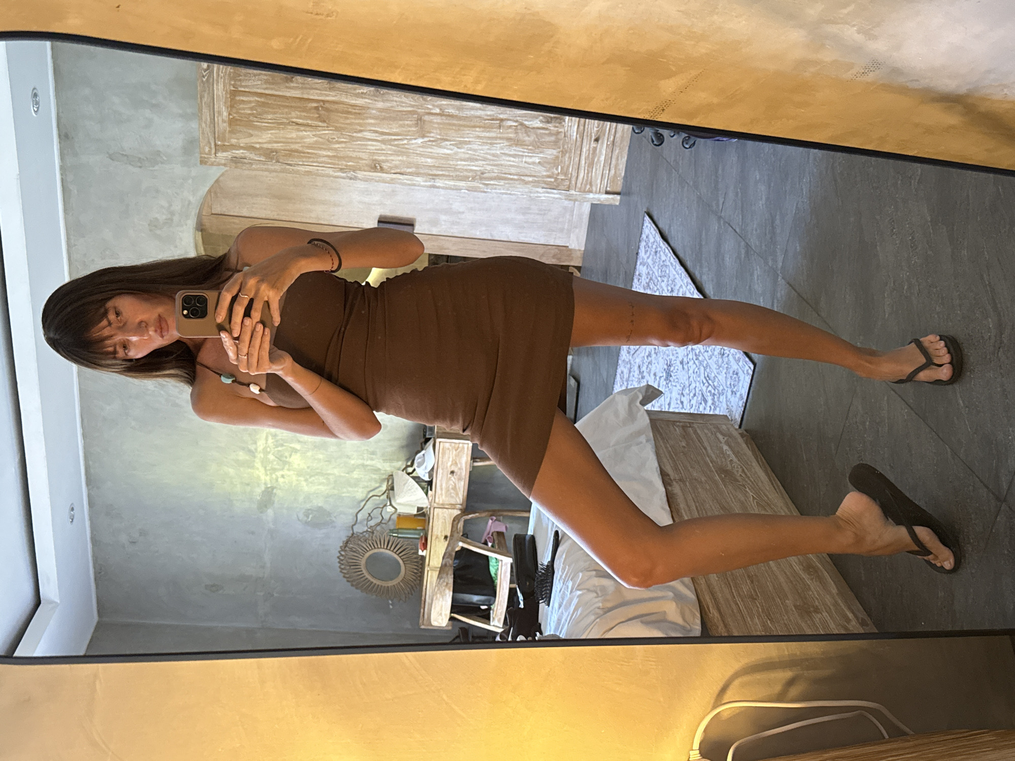 In a bedroom in North Kuta, Indonesia, a woman stands in front of a large mirror, taking a selfie. She is wearing a brown, sleeveless, form-fitting mini dress and dark flip-flops. The room has a tiled floor and a light-colored bed. A wooden dresser with drawers and a mirror is visible in the reflection, as is a wooden wardrobe. The walls are a neutral gray. The lighting suggests it is daytime, with a warm glow coming from a lamp on the left side of the frame.