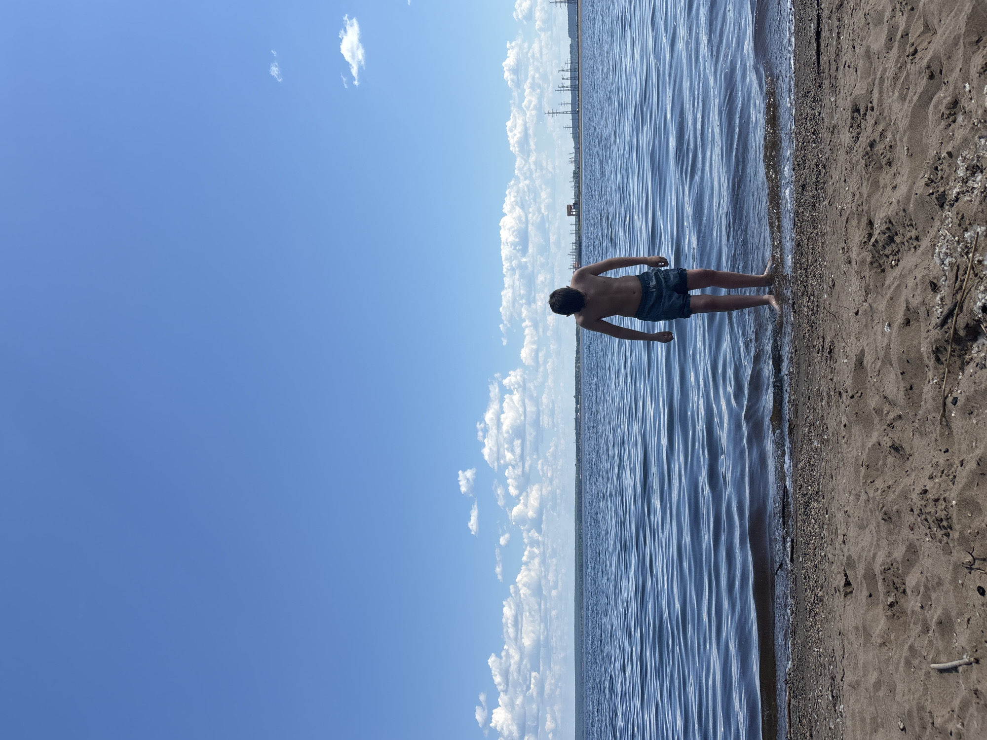 A young male, with dark hair and wearing blue swim trunks, stands at the edge of a body of water. His back is to the camera, and his head is bowed. The water is calm with gentle ripples, reflecting the bright blue sky dotted with white, fluffy clouds. The shore is a mix of sand and pebbles. In the distance, a treeline runs along the horizon, and beyond that, industrial structures with tall transmission towers are visible. The scene appears to be a shoreline of the Kama River in Chaikovsky, Russia, under a clear, sunny sky.
