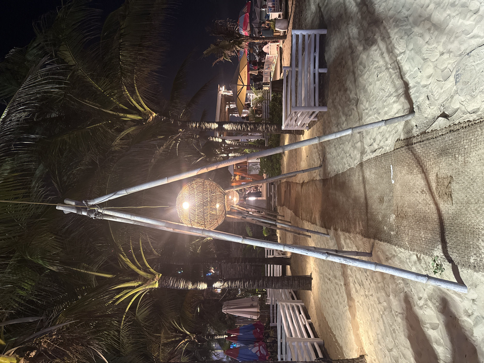 Captured at night, the photograph depicts a beach scene in an unidentified locale within Unknown, Unknown. Several tall palm trees line the left side of the frame. Parallel rows of white benches are situated along the sandy pathway. A series of interconnected, white-painted, triangular structures made of what appears to be bamboo span overhead, each supporting a spherical, intricately woven light fixture. The ground is covered in sand marked with footprints, and a strip of burlap material runs along the right edge of the path. In the background, additional structures and figures, possibly patrons, are discernible under dimly lit conditions. The overall setting implies a relaxed, outdoor dining or recreational space along the beach.