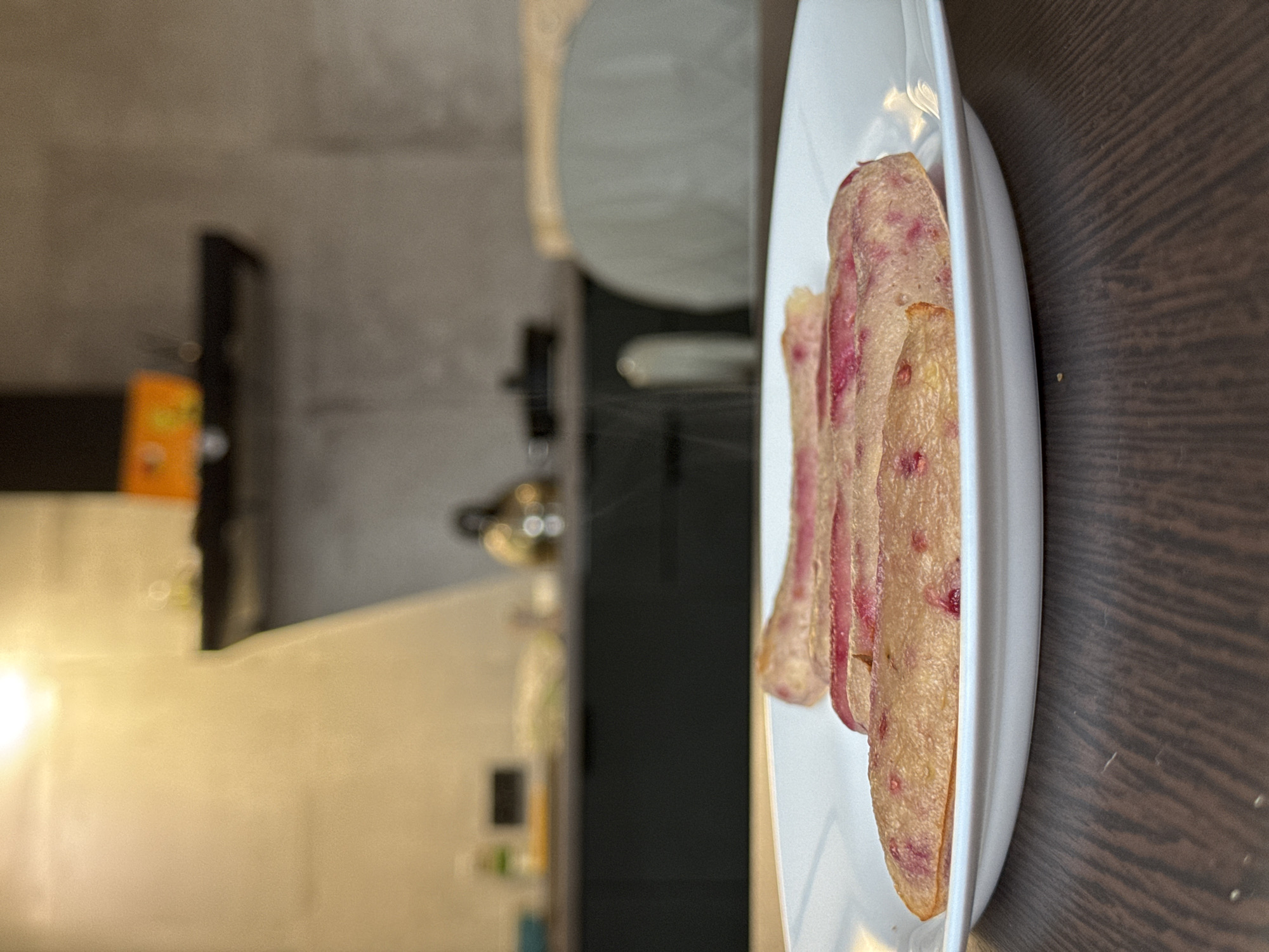 A plate of thinly sliced beet chips sits on a dark wooden surface. In the background, a kitchen is visible, with a kettle and a kitchen cabinet. The location is not provided, and Izhevsk, Russia, cannot be determined from the image.