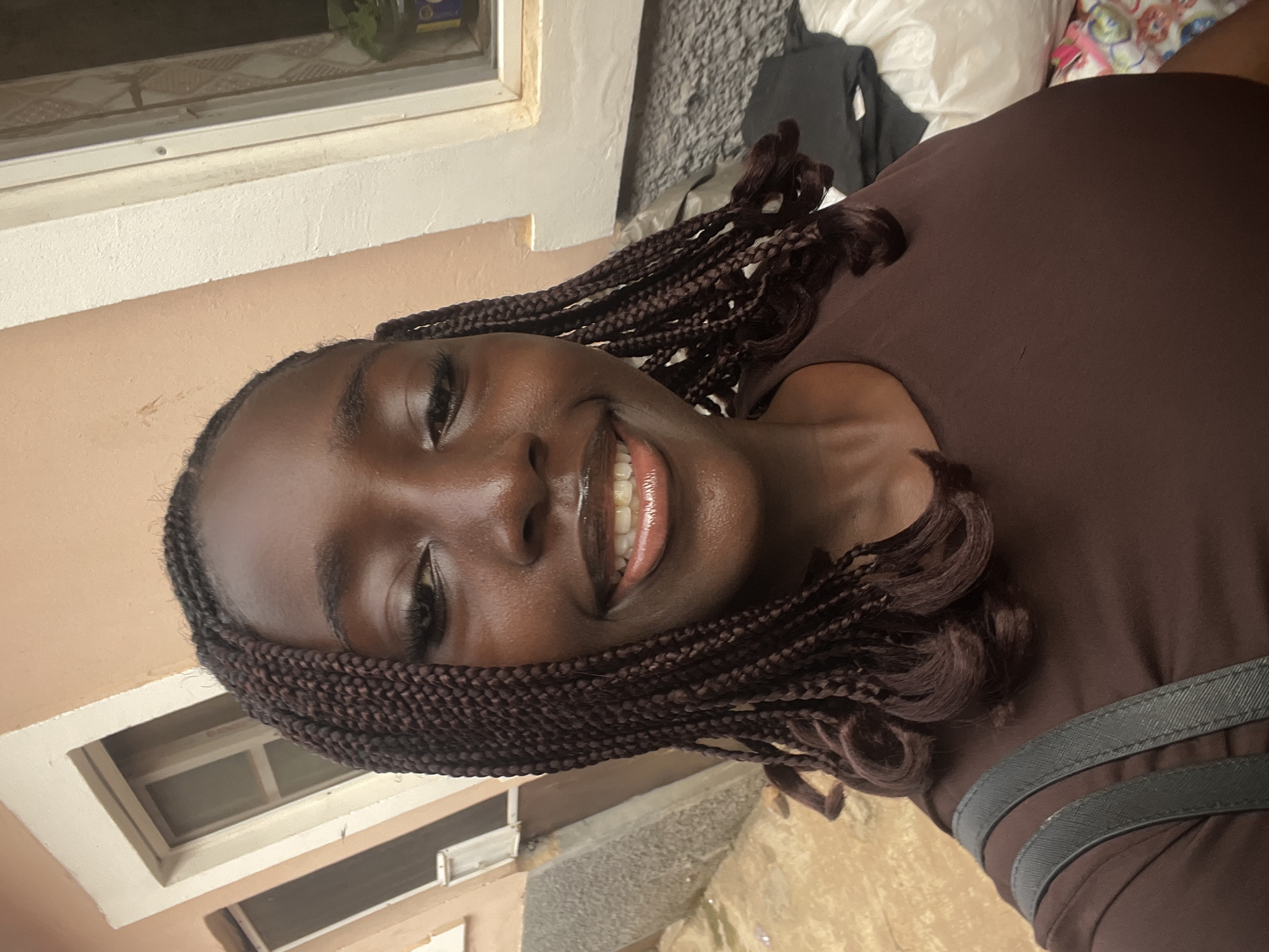 A young woman is depicted outdoors, facing the camera with a smile. She has dark skin and her hair is styled in brown braids. She is wearing a brown top with a strap visible on the right side. The background consists of a beige-colored wall and a window, with a glimpse of foliage and some other items. The location of Abuja, Nigeria cannot be confirmed from the image.