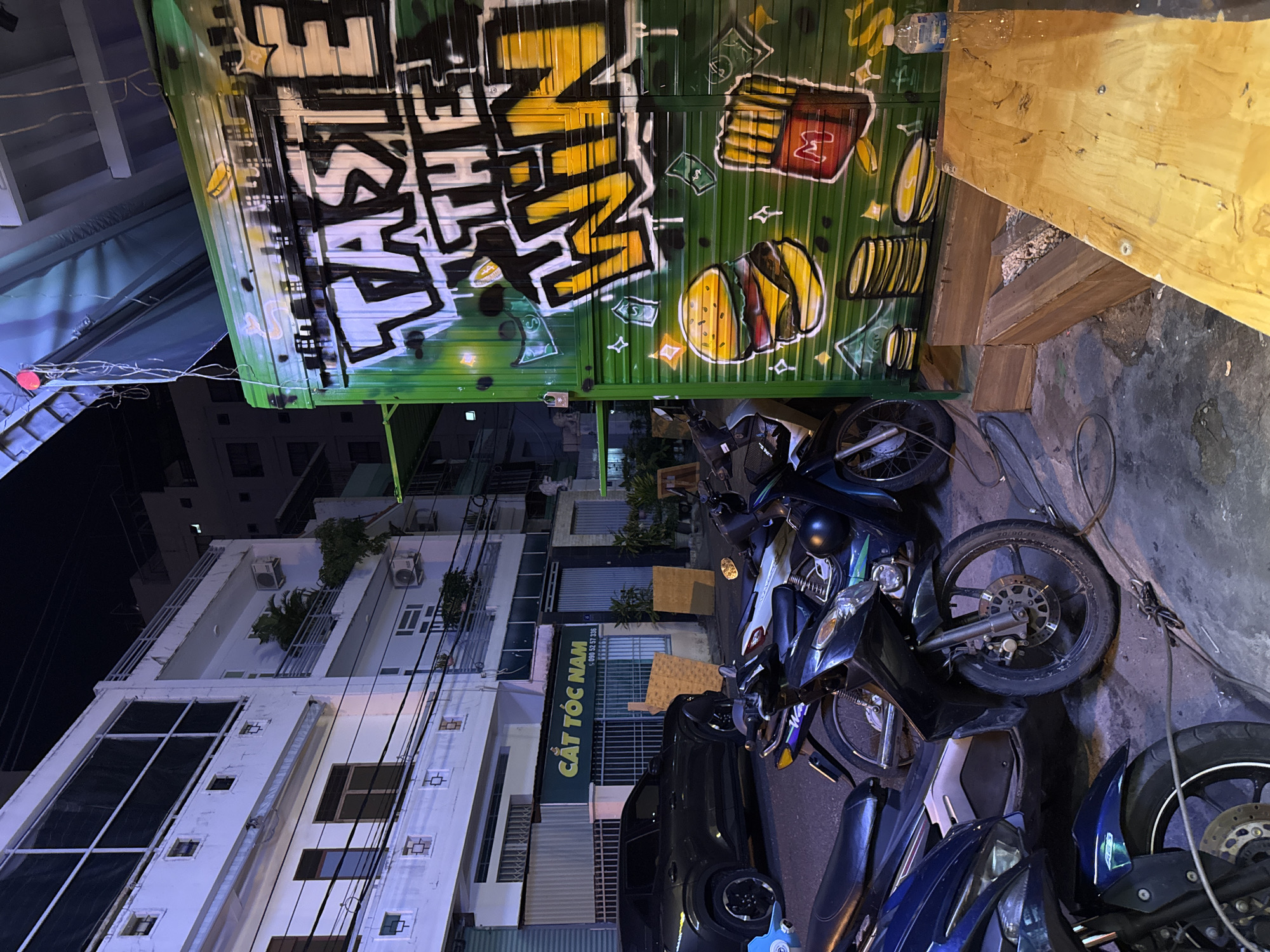 In Nha Trang, Vietnam, at night, a vibrant, graffiti-covered food stall with the words "TASTE THE WIN" is prominently displayed on a green corrugated metal surface. To the left of the stall, a dark-colored SUV is parked on the street. In the foreground, several motorcycles are parked, suggesting a busy urban environment. The buildings in the background are typical of Vietnamese urban architecture, with multiple stories and balconies. The overall scene captures a lively street food culture characteristic of Vietnam.