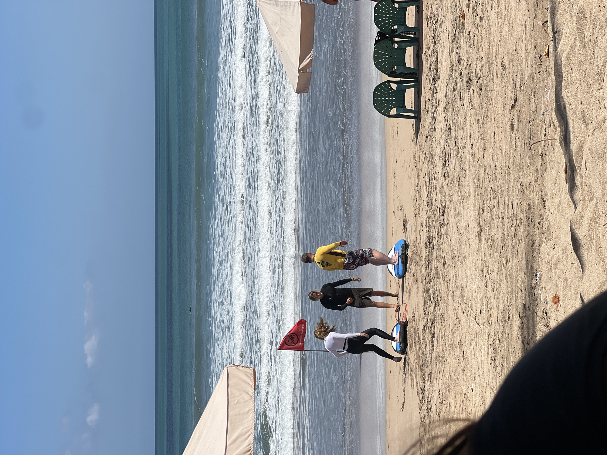 The image depicts a beach scene in Kuta, Indonesia. Three individuals are standing on surfboards on the sandy shore, facing the ocean. A woman in a white long-sleeved top and black leggings is on the left, appearing to be in motion. Next to her is a man in a black long-sleeved shirt and shorts. To the right, a man wears a yellow long-sleeved shirt and patterned shorts. A red flag with "SWIMMING PROHIBITED" written on it is planted in the sand between the woman and the man in black. In the background, the ocean has moderate waves. Two beach umbrellas are visible, one on the left and another on the right, providing shade. Three green plastic chairs are set up on the sand to the right of the frame. The sky is clear and blue with a few scattered clouds. The overall atmosphere is that of a surfing lesson or preparation.