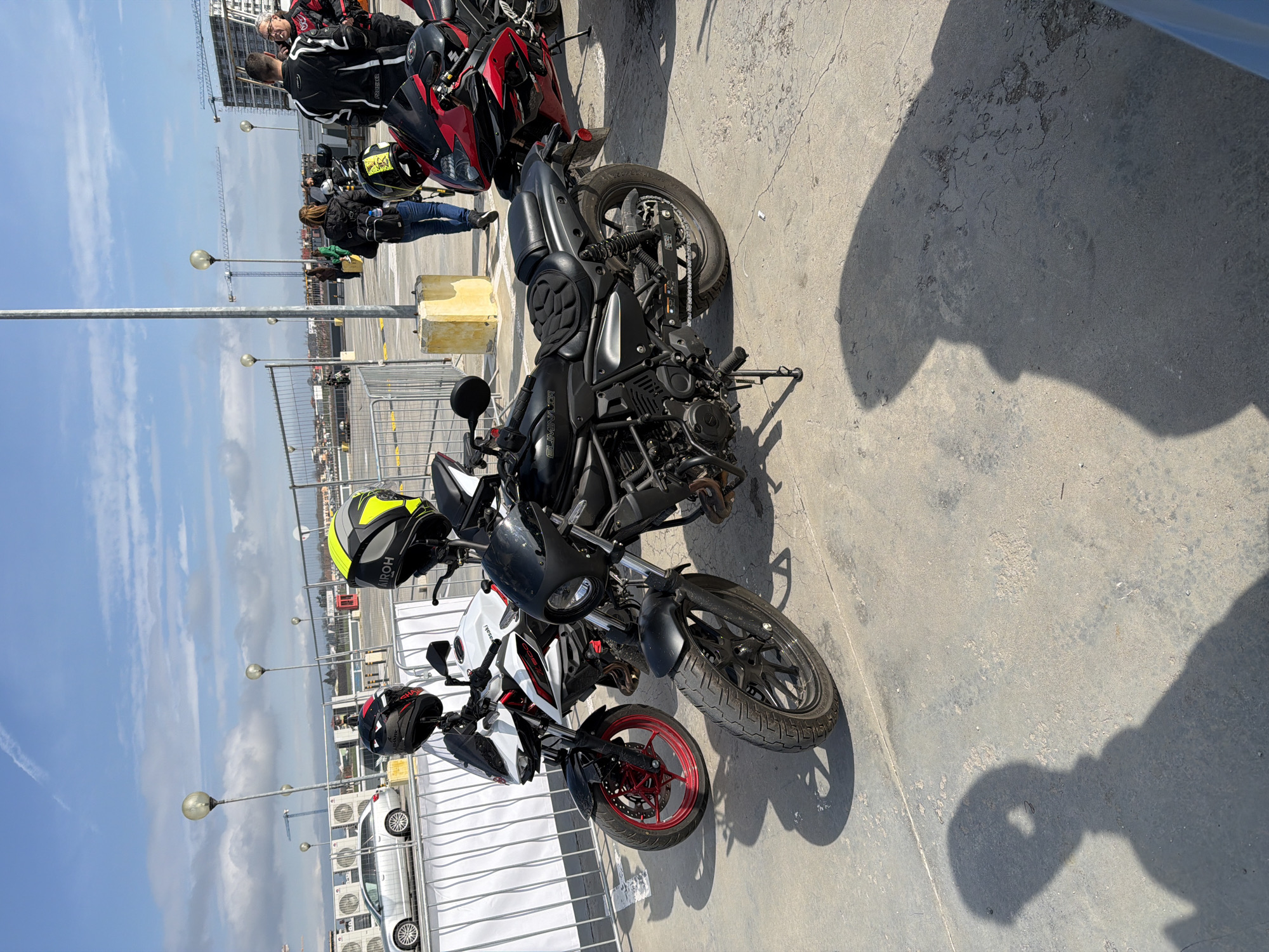 The image shows a group of motorcycles parked on a concrete surface. A white and red Kawasaki motorcycle is positioned near a metal fence, next to a black motorcycle. Helmets are placed on the motorcycles. In the background, there's a glimpse of a cityscape with buildings under construction. People are standing in the distance. Based on the urban elements like the cranes and the specific construction style, the image is likely taken in Burgas, Bulgaria, which is known for its active construction sector and coastal location.