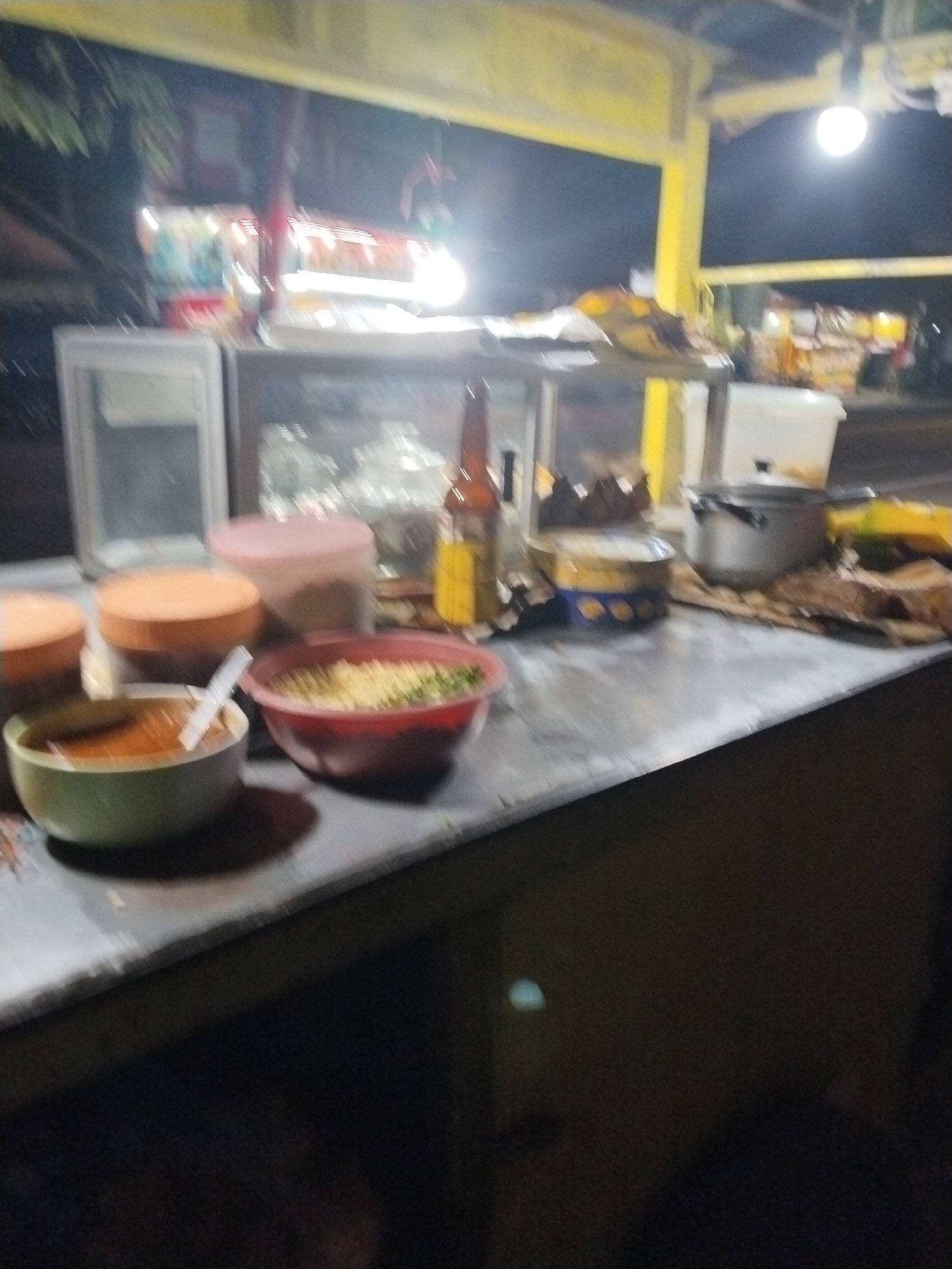 This image depicts an outdoor food stall at night in Suruhan Lor, Indonesia. The stall is illuminated by overhead lights. On the counter, there are several bowls of food, including one with a reddish-orange soup and a white spoon, and another with what appears to be noodles and green vegetables. To the right, there is a stainless steel pot, and behind the counter, various containers and bottles are visible, including a bottle of what looks like soy sauce and a yellow plastic container. The stall appears to be set up along a road, with trees and other structures visible in the background.