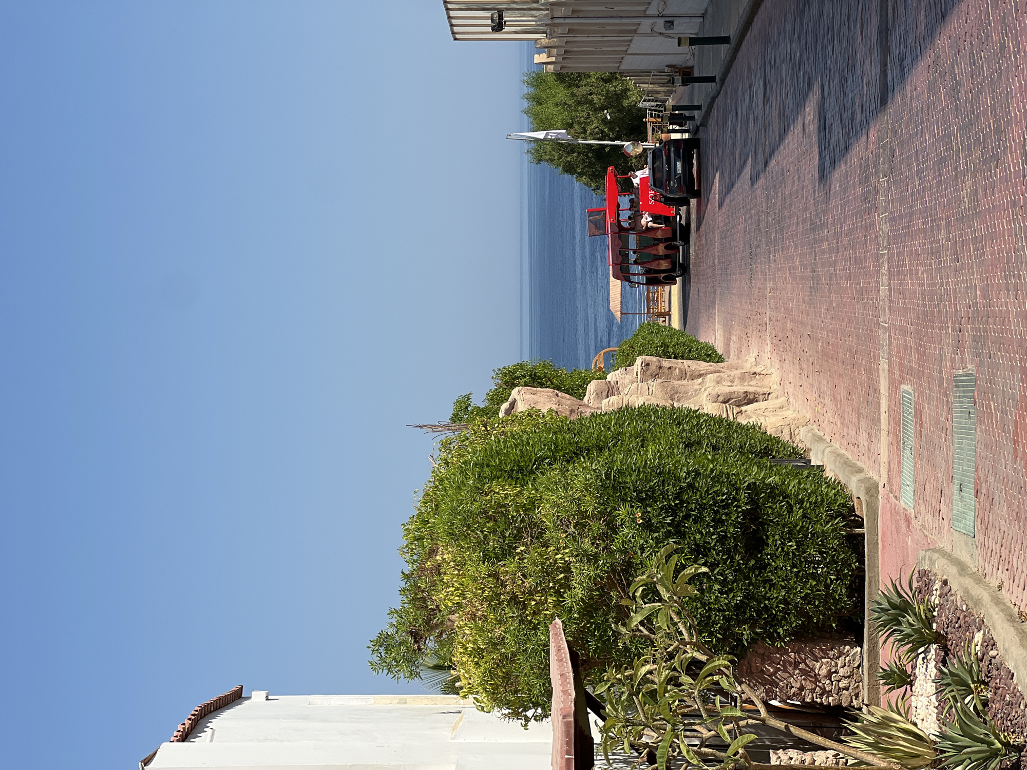 The image depicts a sunlit street in Sharm El Sheikh, Egypt, leading towards the Red Sea. The foreground is dominated by a red brick paved road with a slight incline. To the left, a white building with a terracotta tile roof is partially visible, bordered by lush green foliage and some rocky landscaping. Agave plants are planted in gravel at the base of the foliage.
Further down the road, a red open-air tourist bus carrying passengers is visible. A dark colored SUV follows the bus. Beyond the vehicles, the vibrant blue of the Red Sea stretches to the horizon under a clear, cloudless blue sky. Trees and some structures associated with a beach or resort area are present on the left side of the road near the water. To the right, a building with a vertical striped facade and lights is situated along the sidewalk. Lampposts line the right side of the road. The scene conveys a typical resort atmosphere, with elements of architecture, nature, and transportation common to a tourist destination in Sharm El Sheikh.
