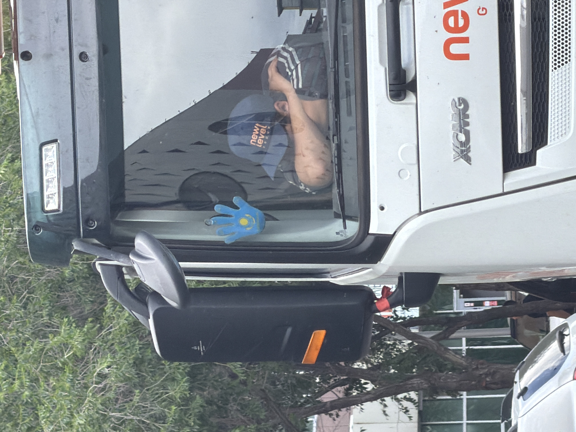 The image shows the exterior of a white truck, likely a commercial vehicle, parked on a street. A person is asleep in the driver's seat, their head resting on their arms and leaning against the windshield. They are wearing a blue baseball cap with the text "new level" and an athletic jacket with three stripes. A blue, hand-shaped air freshener with a yellow sun and star emblem, resembling the flag of Kazakhstan, is hanging from the rearview mirror. The truck has a side mirror with an orange reflector and a red ribbon tied to its support arm. The truck's grill has "XCMG" and "new G" branding visible. The background consists of trees and buildings, suggesting an urban or semi-urban setting. The overall scene depicts a moment of rest for the driver in what appears to be the city of Astana, Kazakhstan.