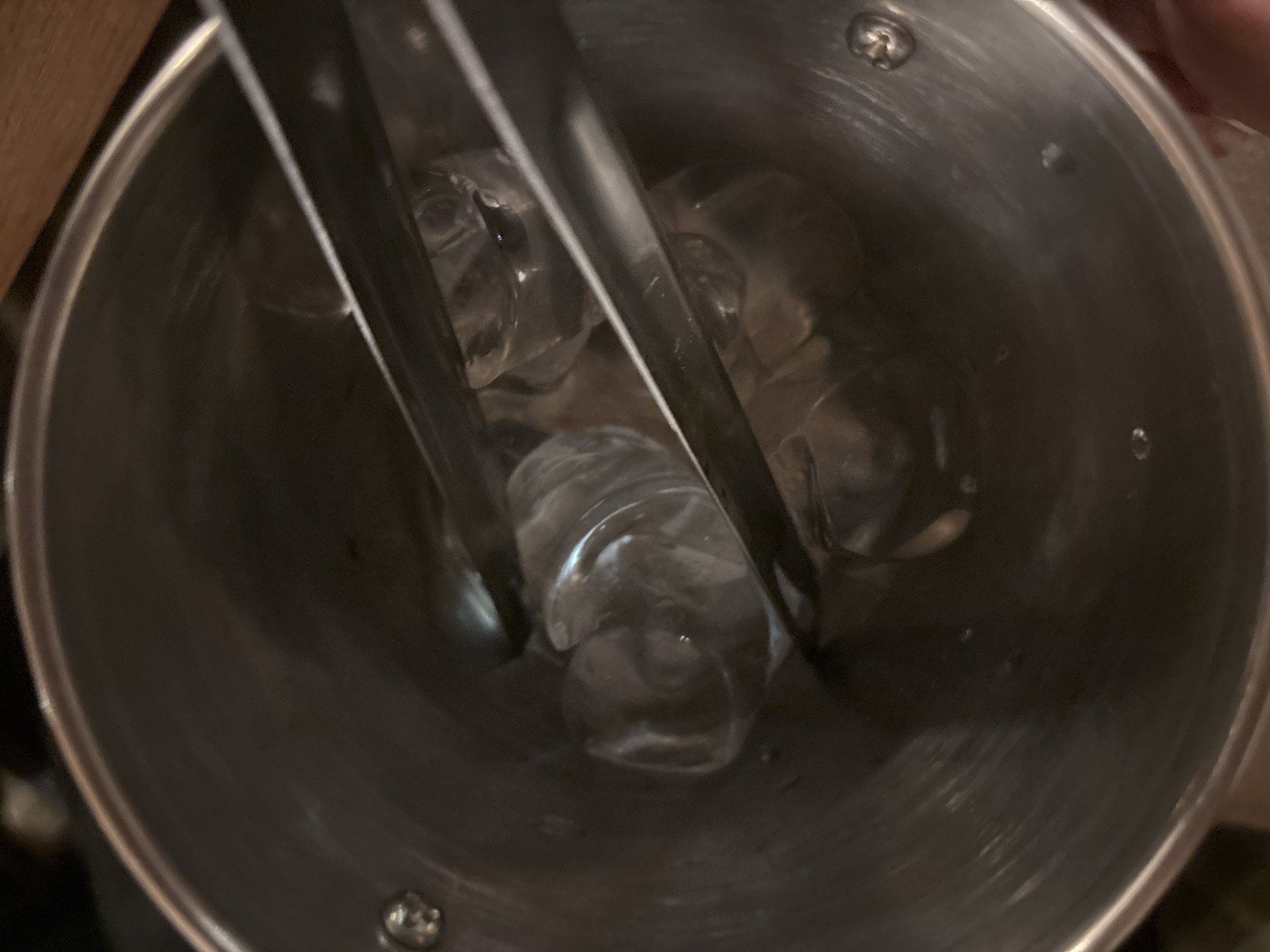 A top-down close-up shot shows ice tongs holding ice cubes inside a metal bucket. The bucket's interior is visible, revealing several ice cubes. The bucket has a metallic finish. The background is out of focus, only revealing a glimpse of a wooden surface and dark, indistinct elements. No information is available to determine the location in Sofia, Bulgaria, or any information about the presence or absence of people.