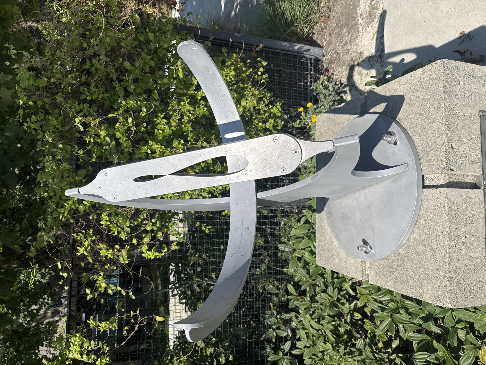 The image displays a modern, metallic sundial sculpture situated outdoors on a concrete pedestal. The sculpture has a curved, arc-like structure with markings indicating hours and possibly months. It is positioned in a landscaped area with lush green foliage and a chain-link fence visible in the background, suggesting an urban or park-like setting. The sunlight casts distinct shadows from the sculpture onto the ground. The sculpture is located at the Burnaby Public Library, specifically outside the McGill Library branch.