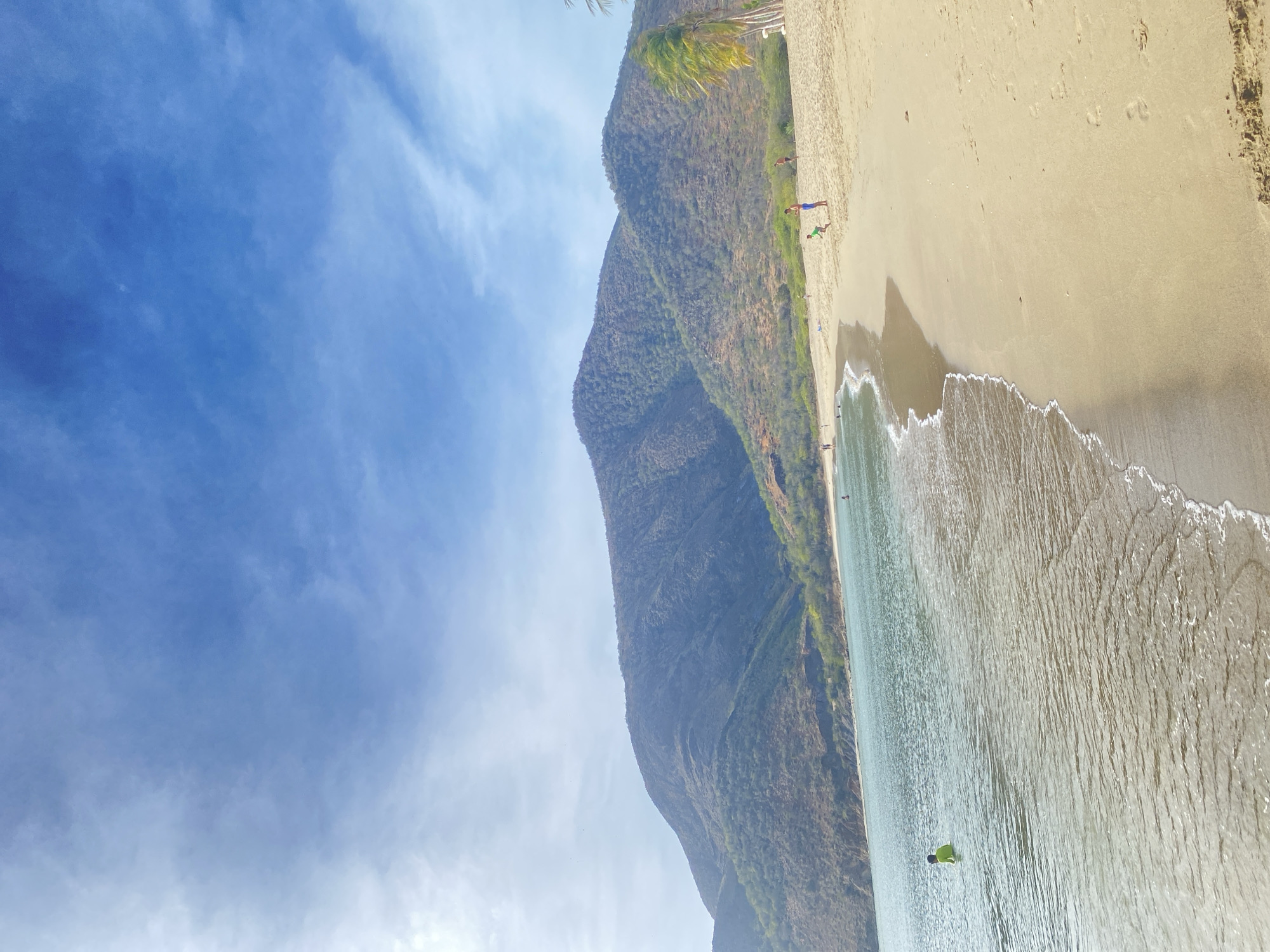 The image depicts a beach scene in Juan Griego, Venezuela. A large, green-covered mountain slopes down towards the beach. A sandy beach is visible with gentle waves breaking along the shoreline. Several people are visible on the beach and in the water. The sky is blue with scattered clouds.