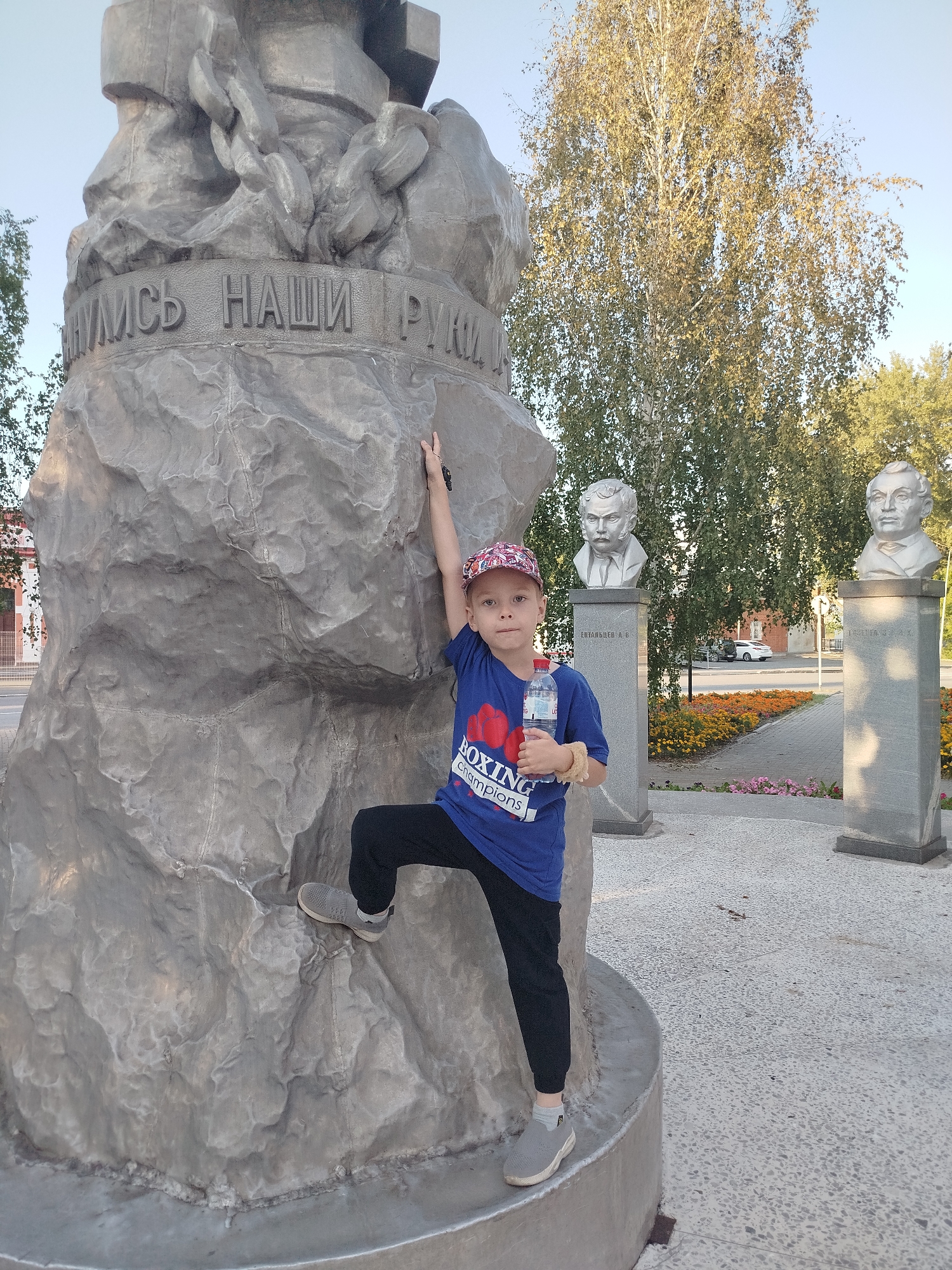 The image shows a young boy climbing on a large monument with carved text in Russian, located in the city of Yalutorovsk, Russia. The monument appears to be made of stone and features reliefs of hands and chains. The boy is wearing a blue t-shirt with graphics, black pants, and a cap. He is holding a bottle of water. In the background, there are busts of two men on pedestals, a tree with yellowing leaves, and a street with some traffic and buildings. The ground is paved with small stones. The overall scene suggests an outdoor public space in a city.